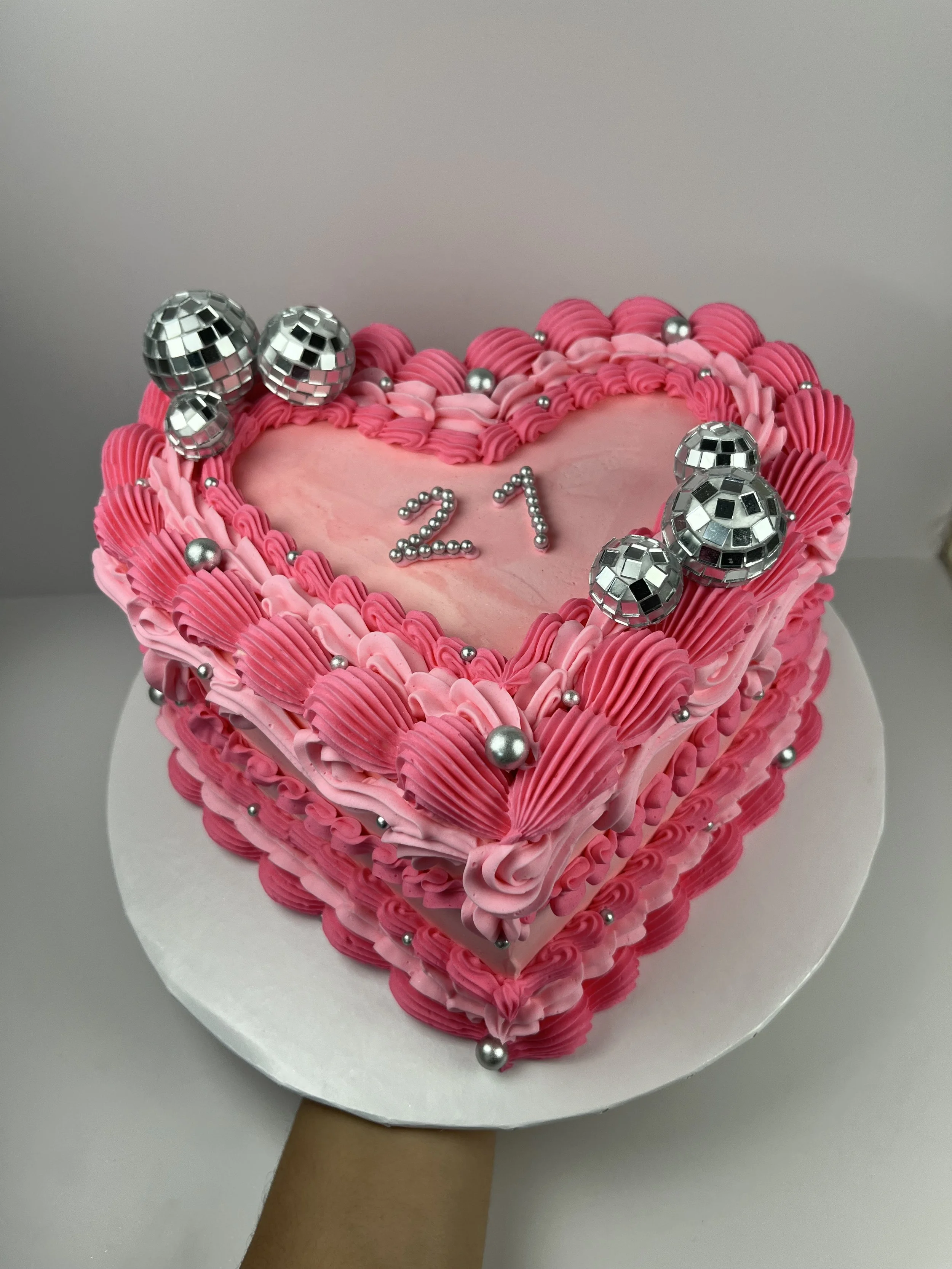 Pink heart-shaped cake decorated with pink and silver accents, including edible pearls and disco ball decorations, with '21' written on top, held on a white cake board.