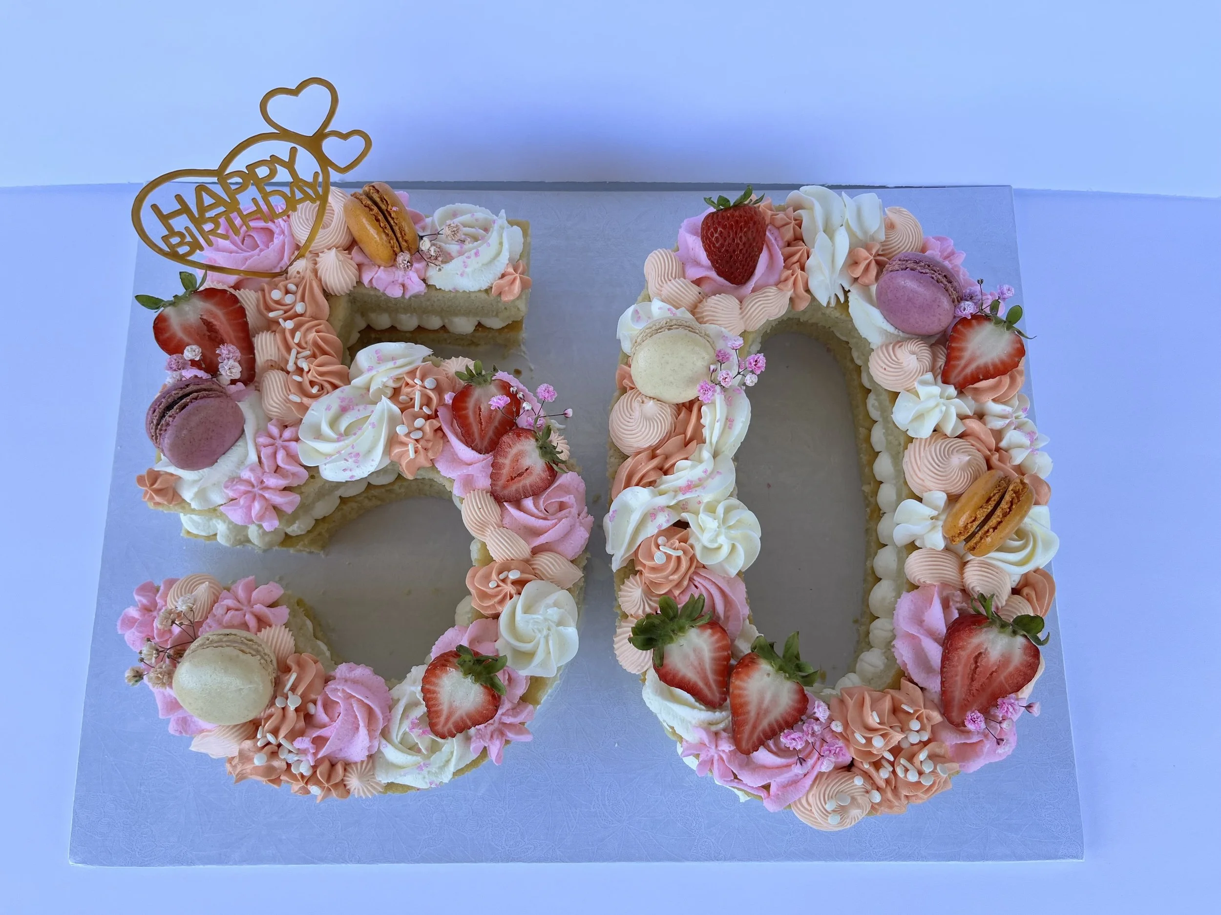 A birthday cake in the shape of the number 20, decorated with pink and white icing, strawberries, macarons, macarons, and small flowers. A cake topper reads 'Happy Birthday' with two heart-shaped decorations.