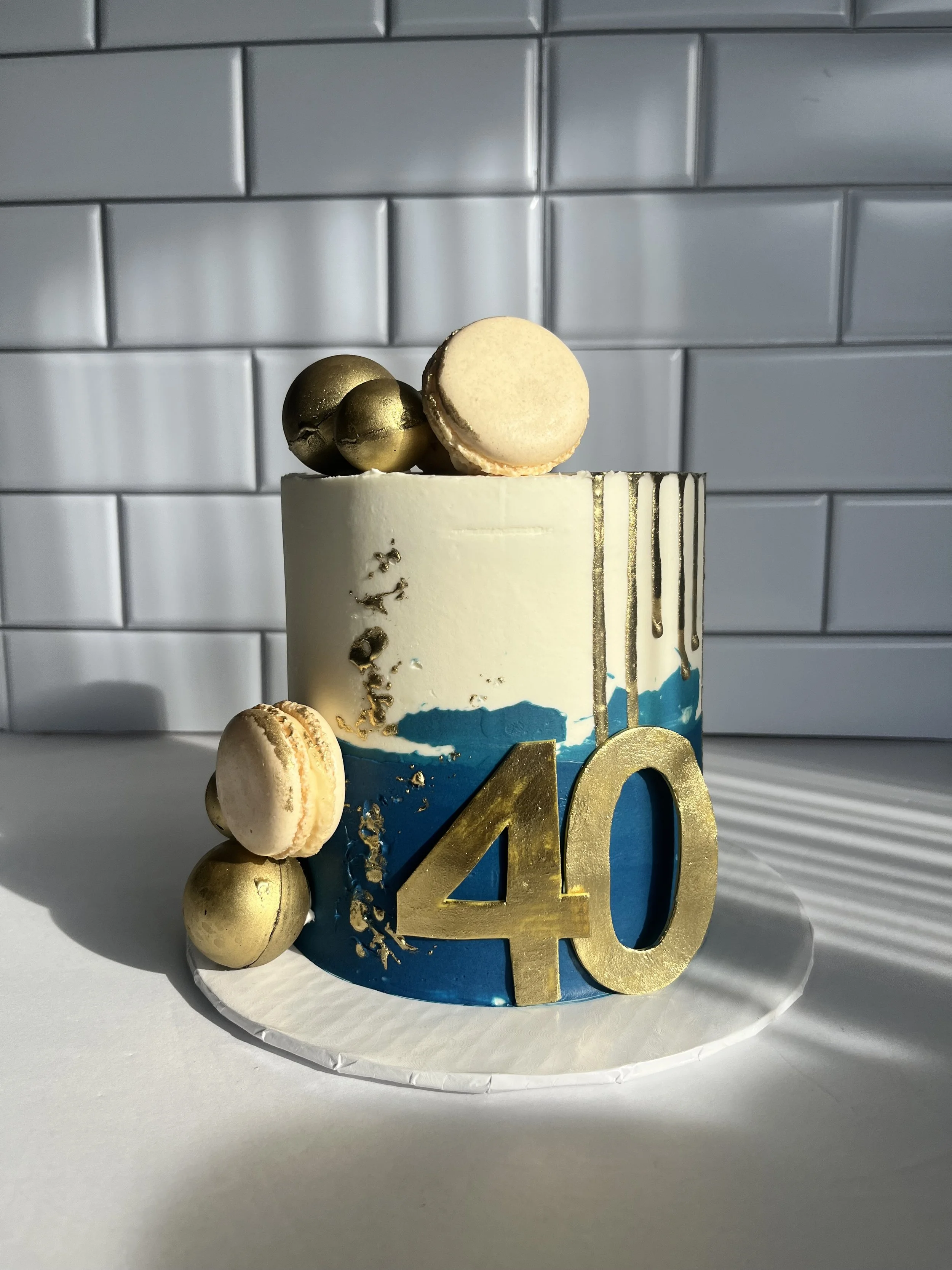 A cake decorated with gold spheres, macarons, and a large gold '40' topper, set against a tiled gray background.