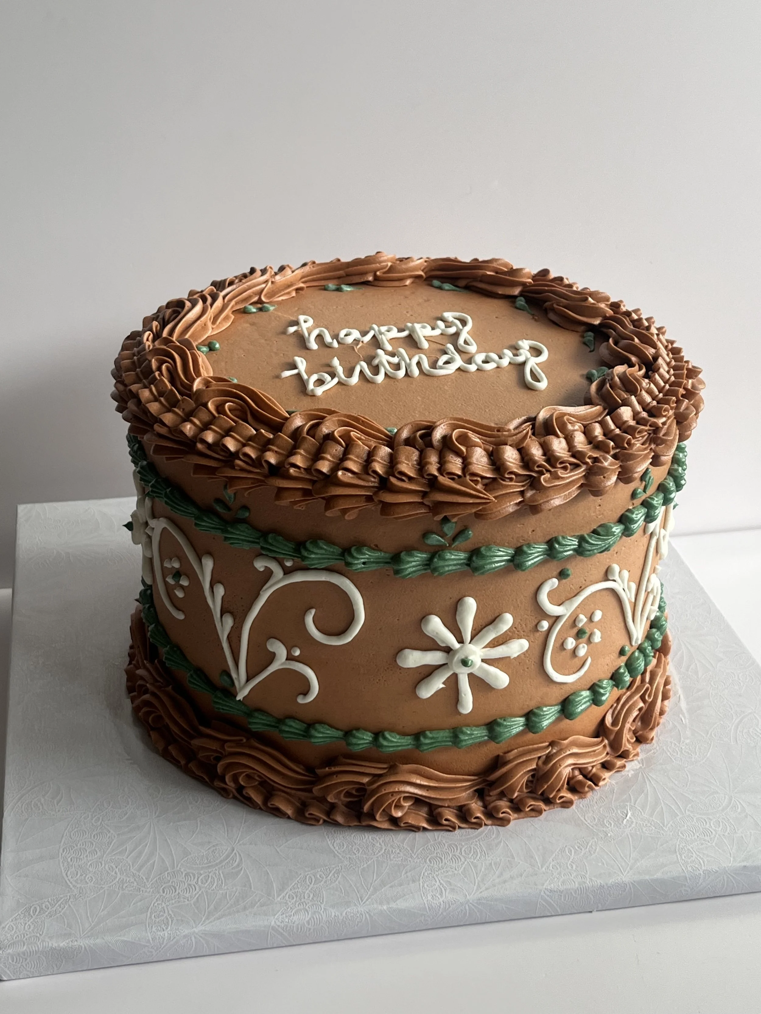 Two-tier brown birthday cake decorated with green and white icing, with 'happy birthday' written in white icing on top.
