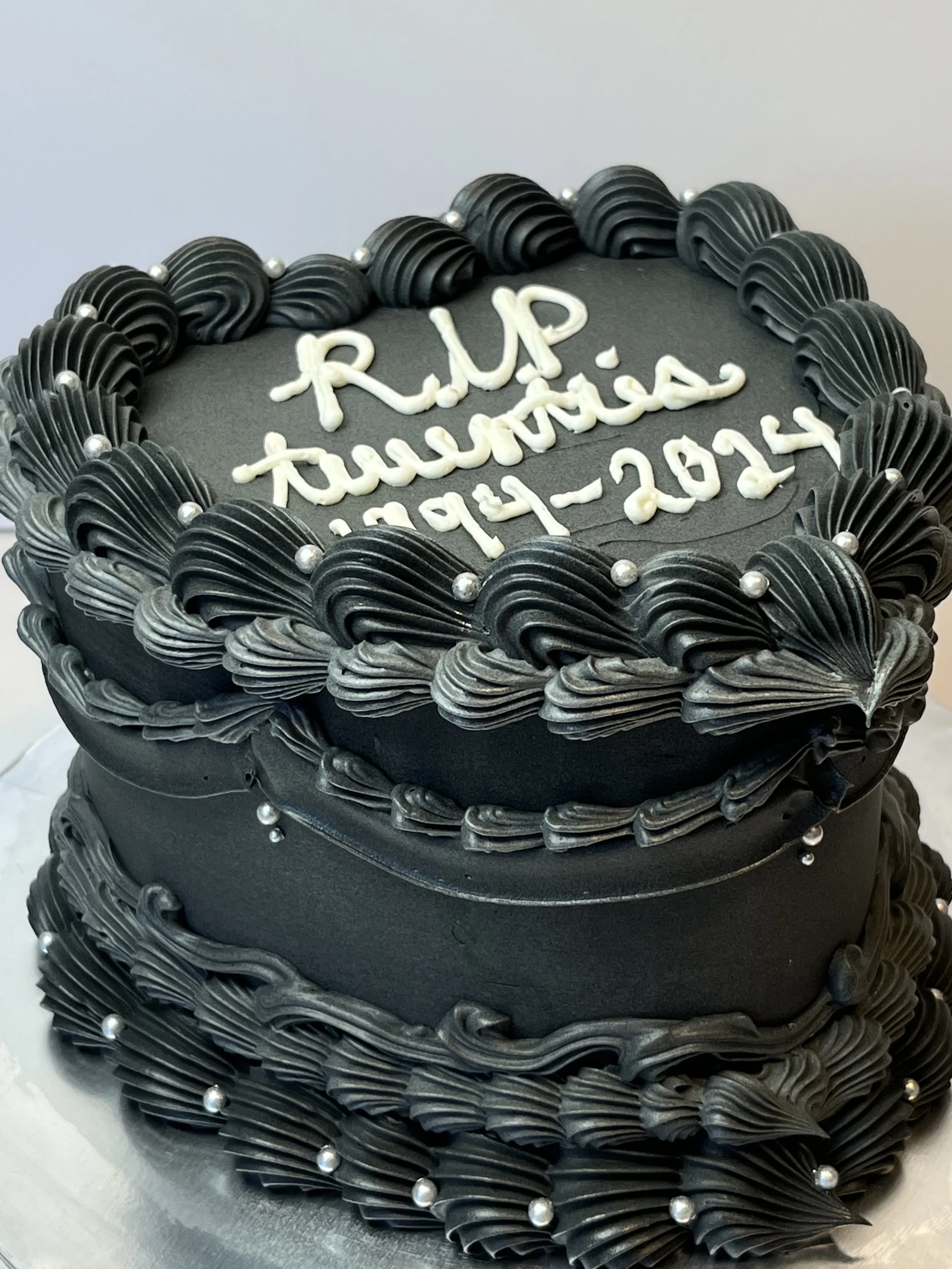 A black cake decorated with black frosting, silver edible beads, and white writing that says 'RIP Trusted' along with the date '11-21-22'.
