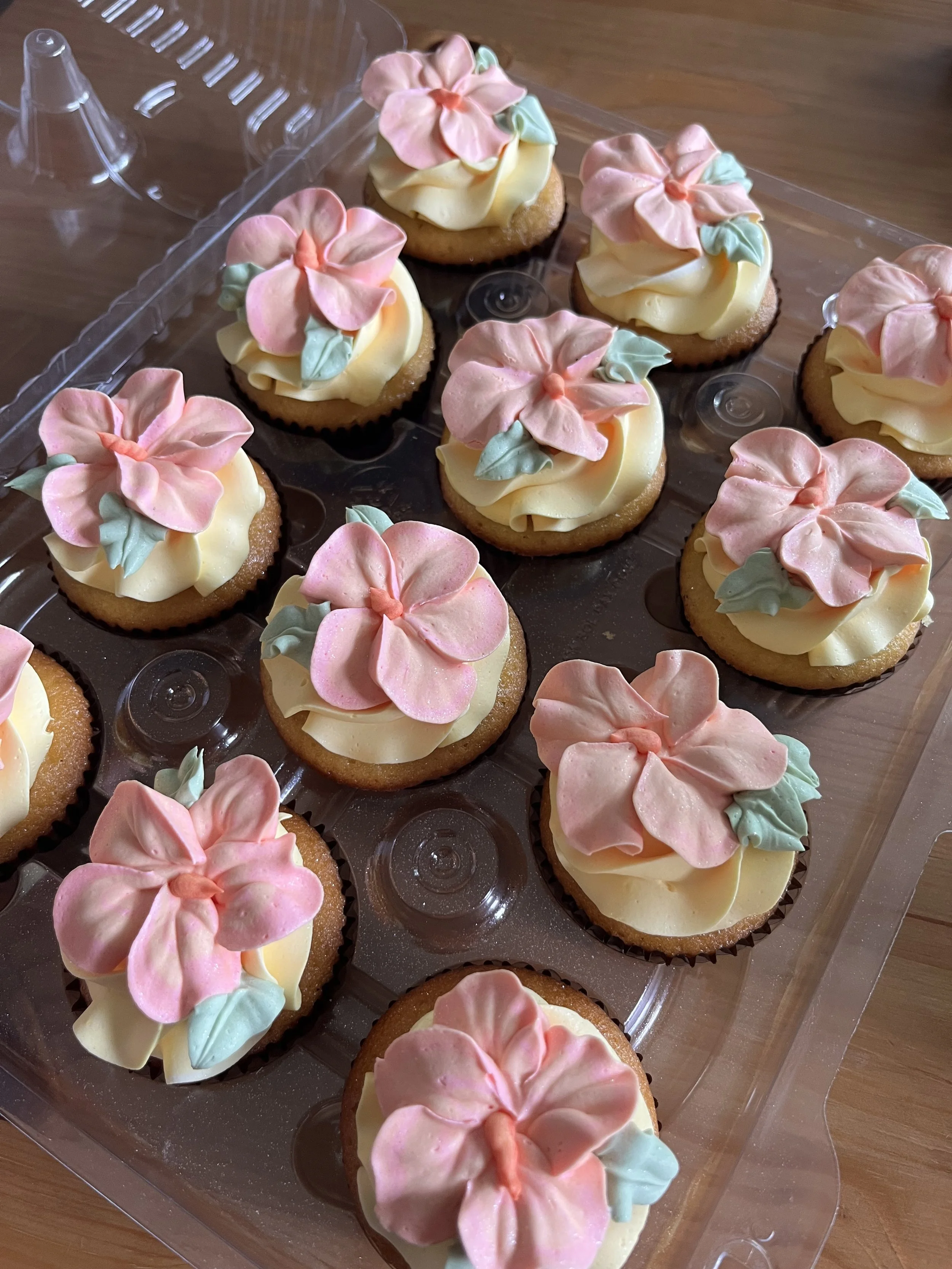 A plastic container filled with cupcakes decorated with pastel-colored buttercream flowers.