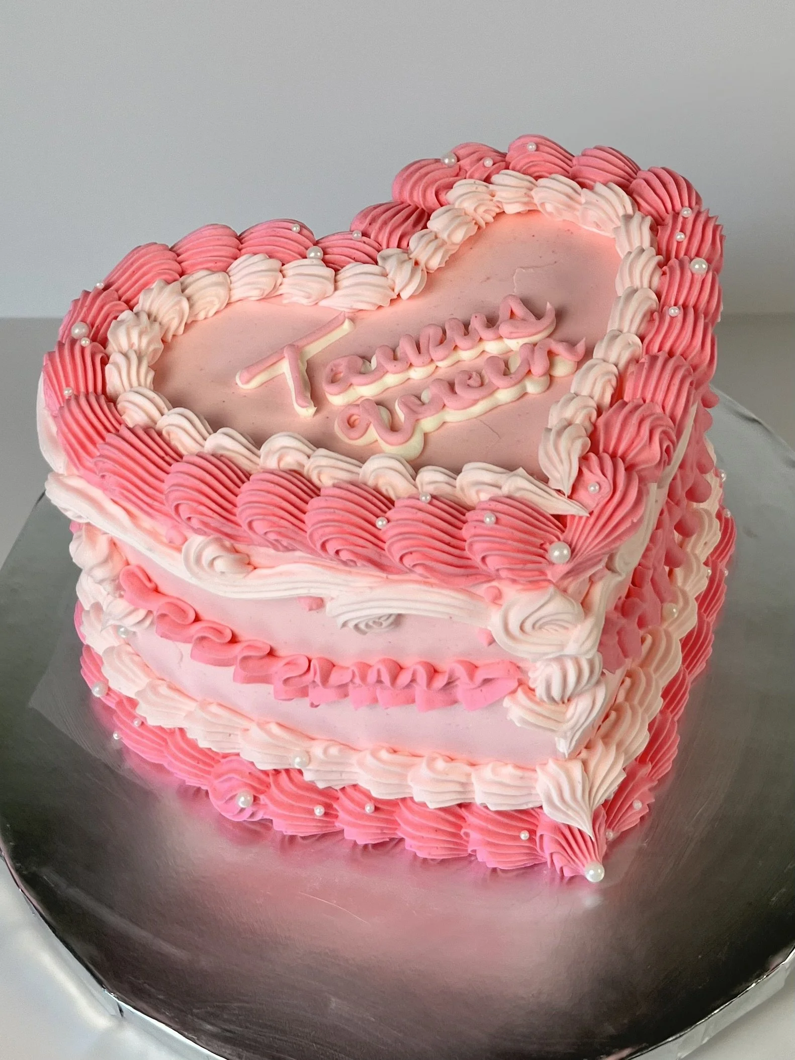 A pink heart-shaped cake decorated with pink and white piped frosting and small edible pearls, with the words 'Te quiero' written in pink icing on top.
