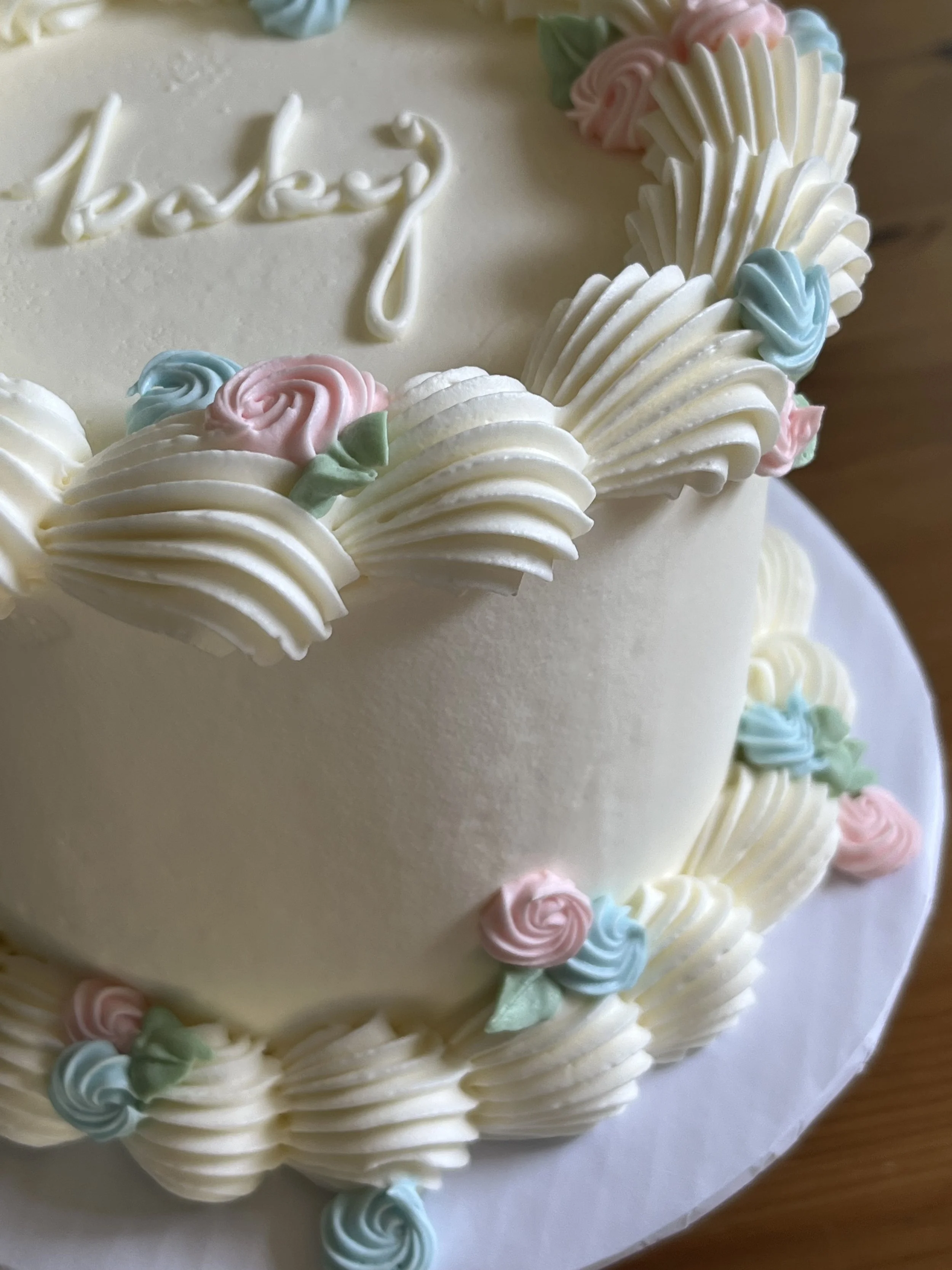 Close-up of a decorated birthday cake with white frosting, pastel pink, blue, and green frosting roses, and a white cursive message on top.