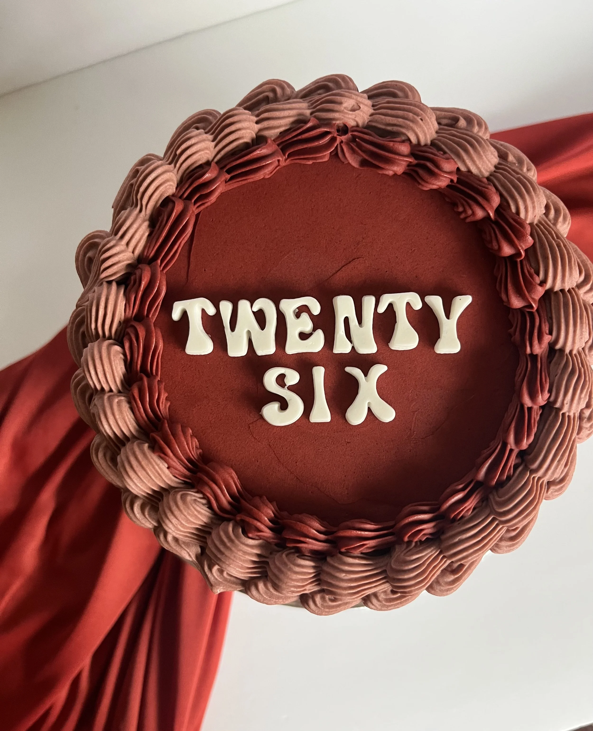 close-up of a red heart-shaped cake with pink and red piped icing border, white fondant letters reading 'TWEETY', and red satin ribbon decoration, with a plain white background and red drapery.