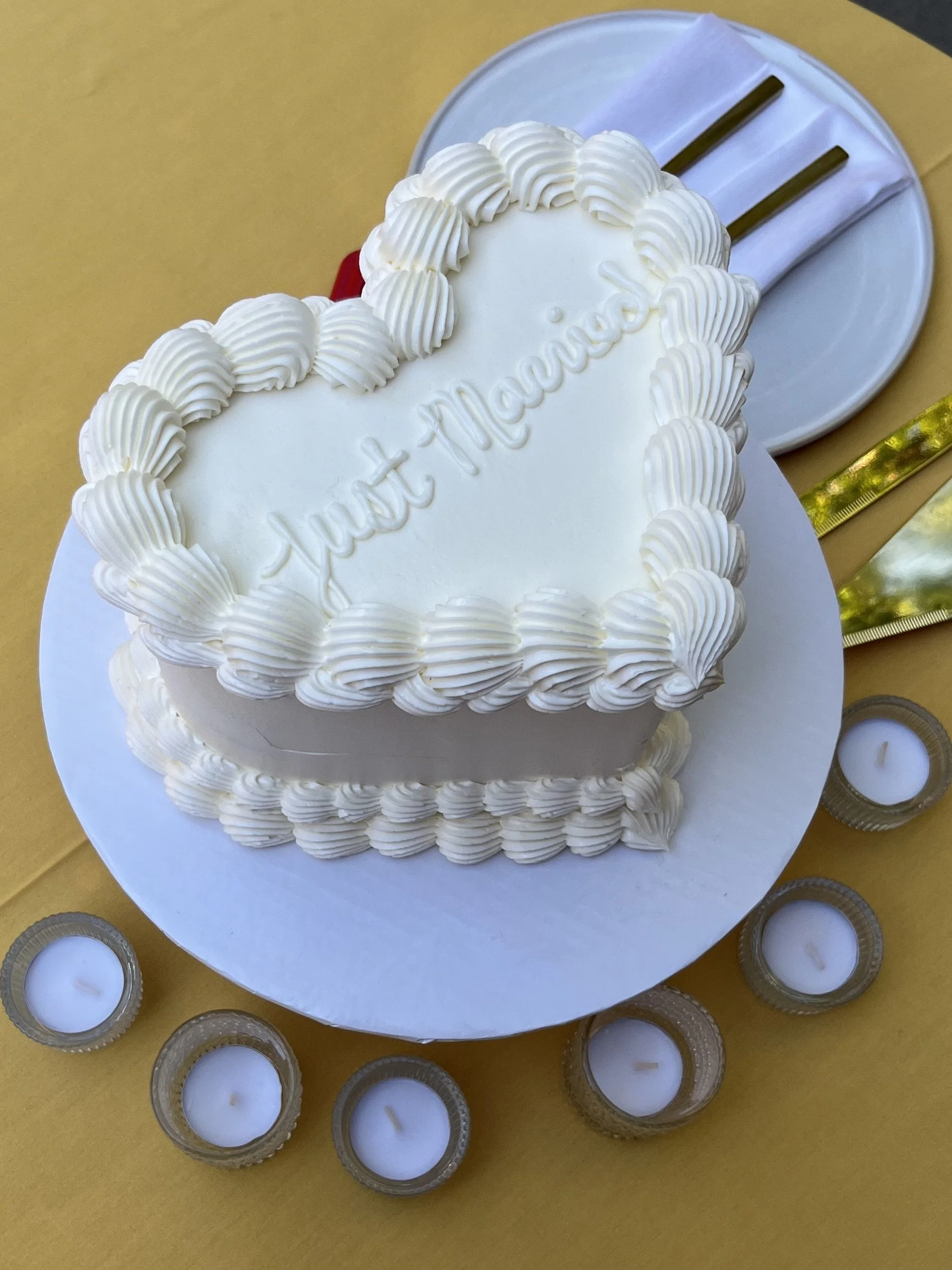 White heart-shaped cake with piped cream border and the words "Love Marwed" on top, surrounded by small tea light candles on a yellow table.