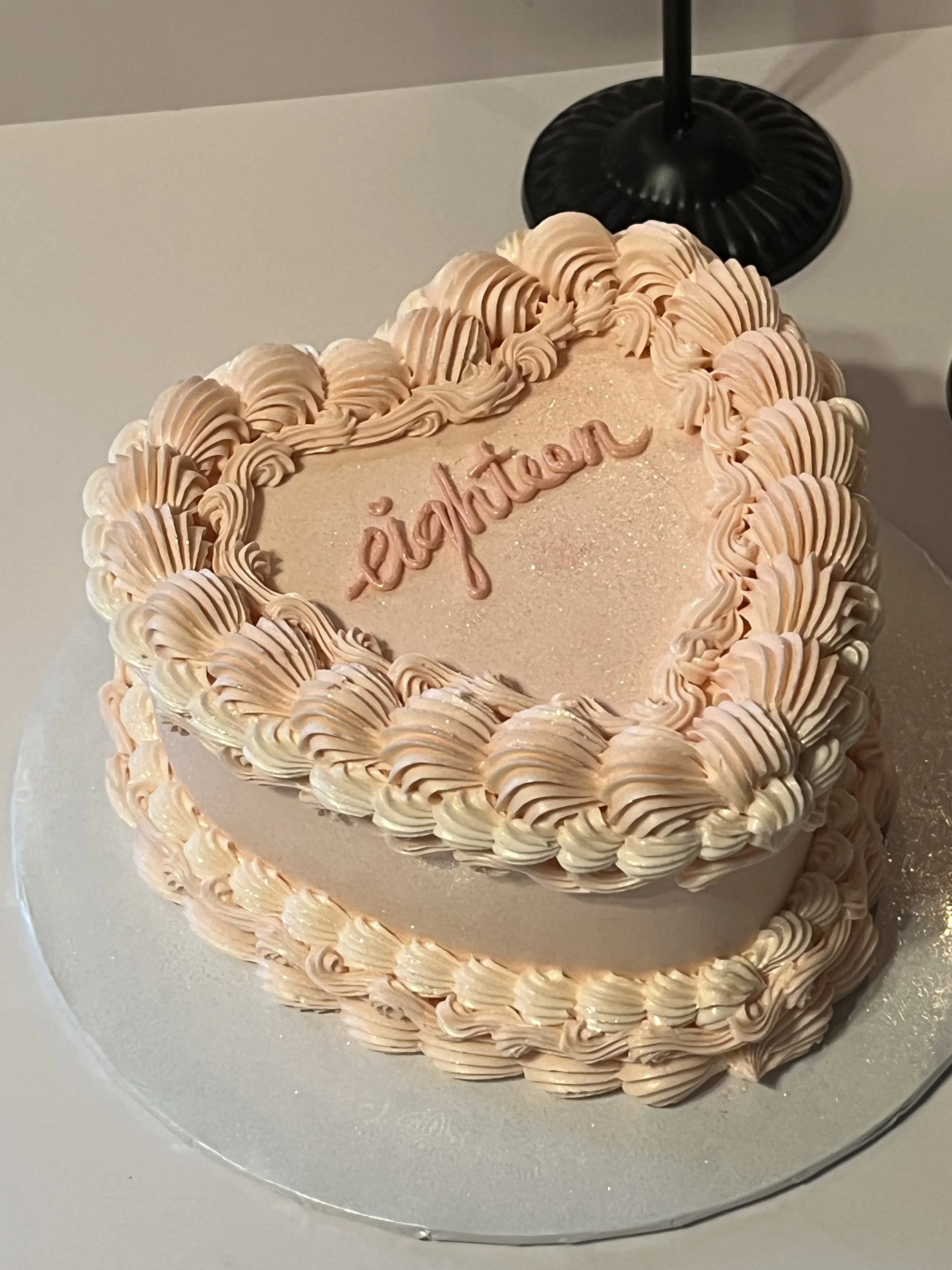 A round birthday cake decorated with peach-colored frosting, piped in ruffled and swirl patterns, with the name 'Eighteen' written on it.