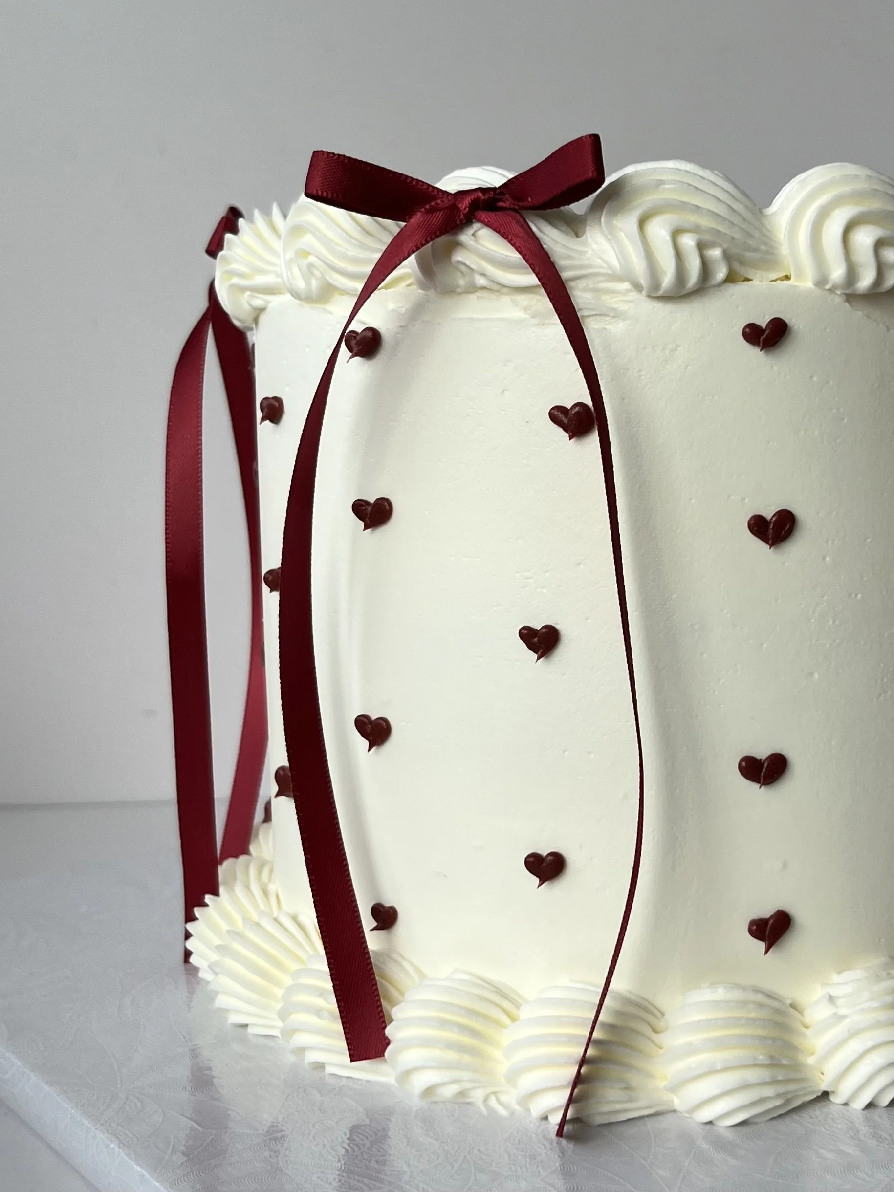 A white wedding cake decorated with a burgundy bow, burgundy heart-shaped sprinkles, and cream-colored piped frosting on the top and bottom edges.