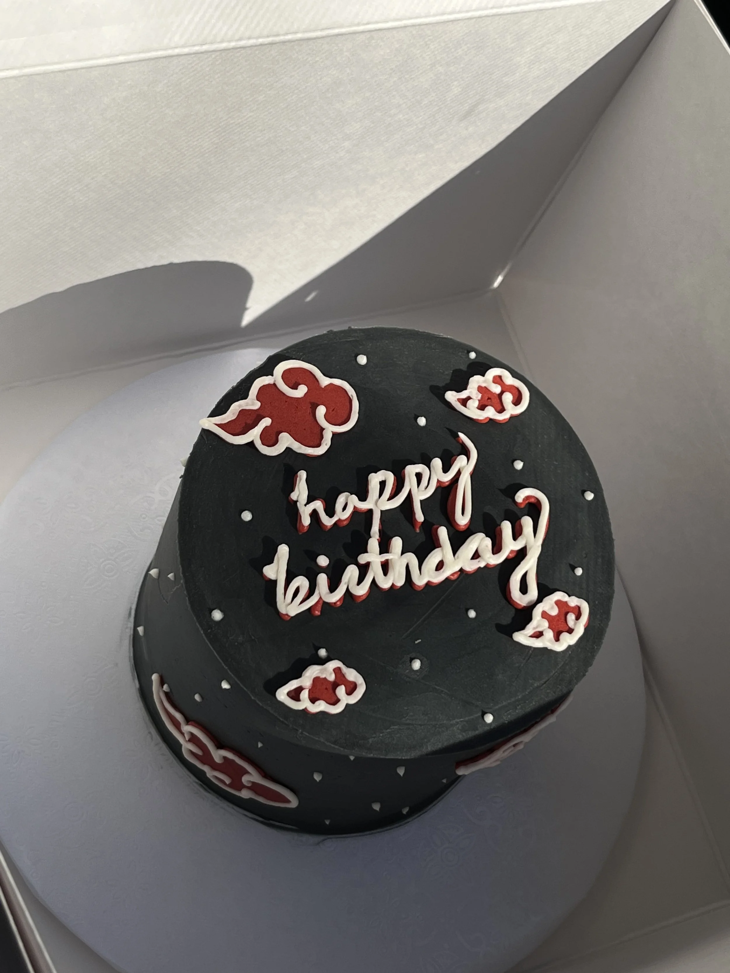 A black birthday cake with red and white decorations, topped with a message saying 'happy birthday' and surrounded by red and white floral designs, inside a white cake box.