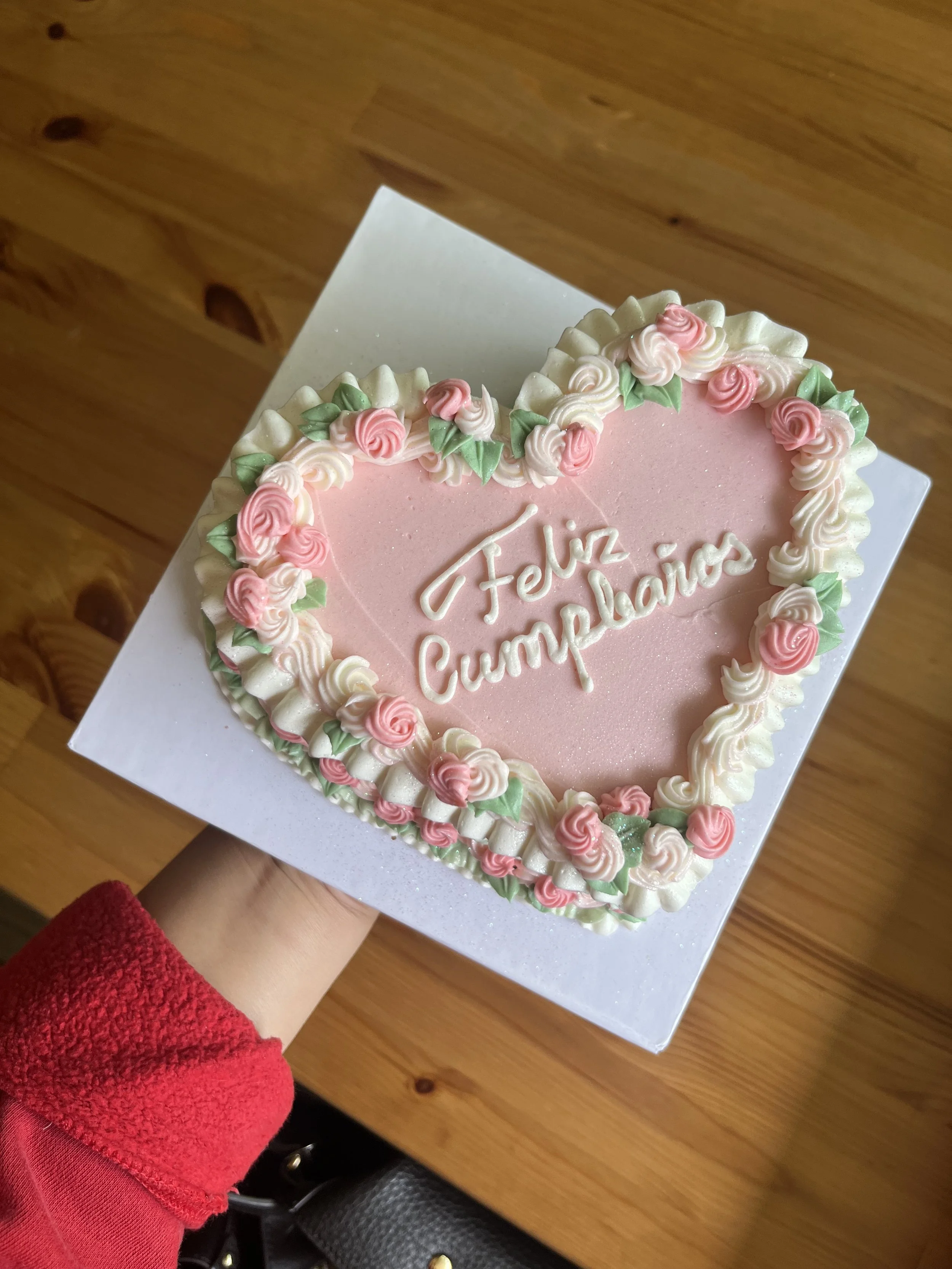 Pink heart-shaped cake with floral decorations and the words 'Feliz Cumpleaños' written in white icing, being held by a person in a red sweater over a wooden table.