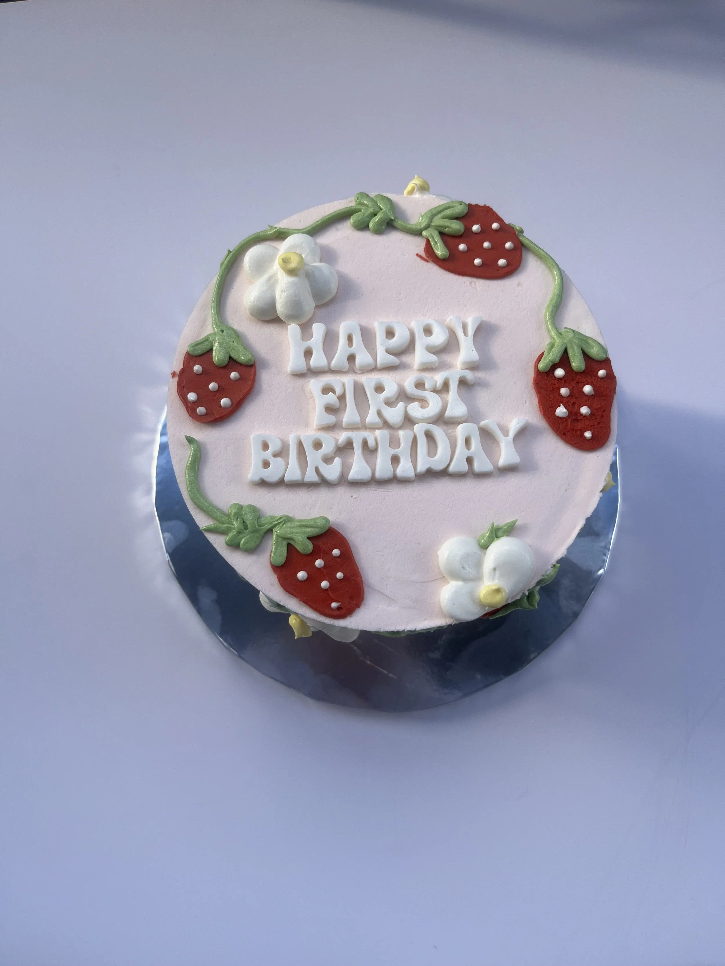 A round birthday cake with pink icing decorated with strawberry and white flower designs, featuring white letters spelling 'Happy First Birthday.'