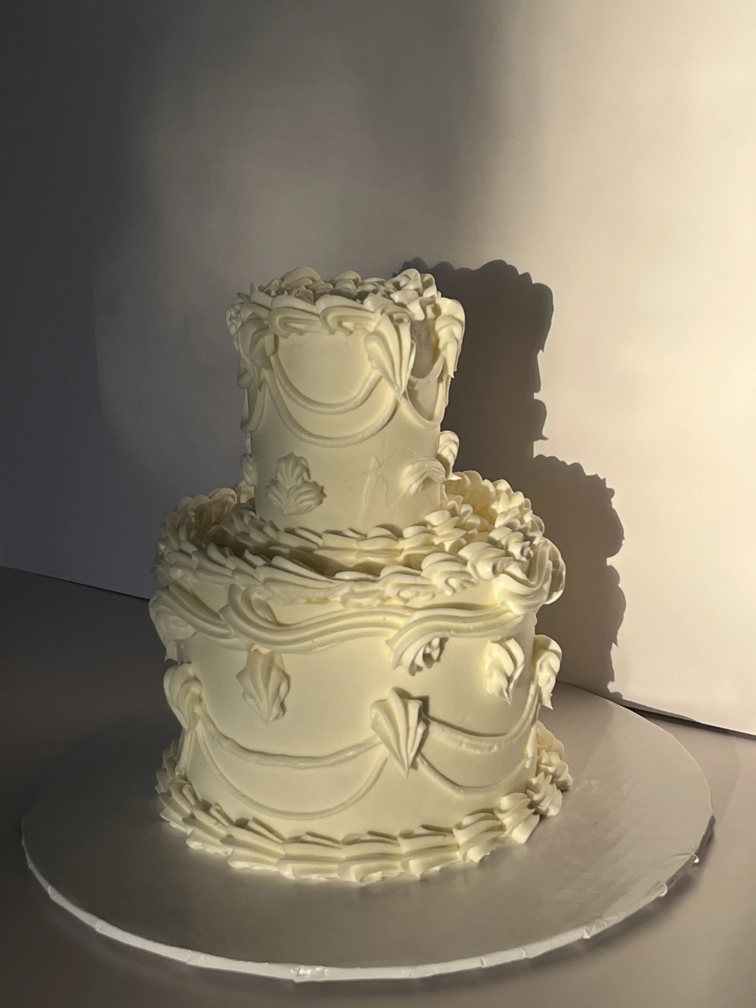 Decorative cream-colored tiered cake with intricate piped floral and scroll designs, displayed on a silver cake board.