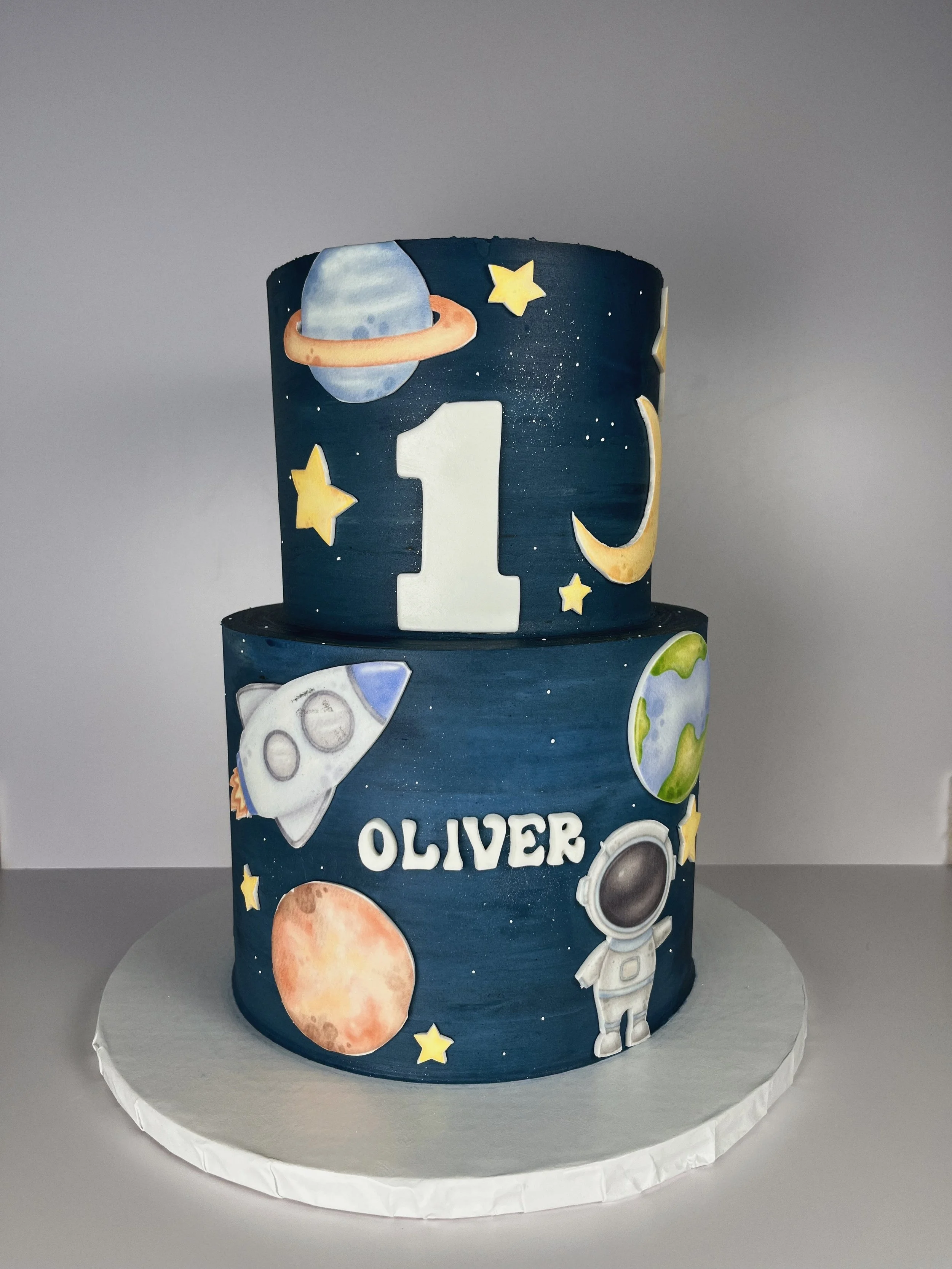 A space-themed birthday cake with the name 'Oliver' written on it, decorated with planets, stars, rockets, an astronaut, and space illustrations.