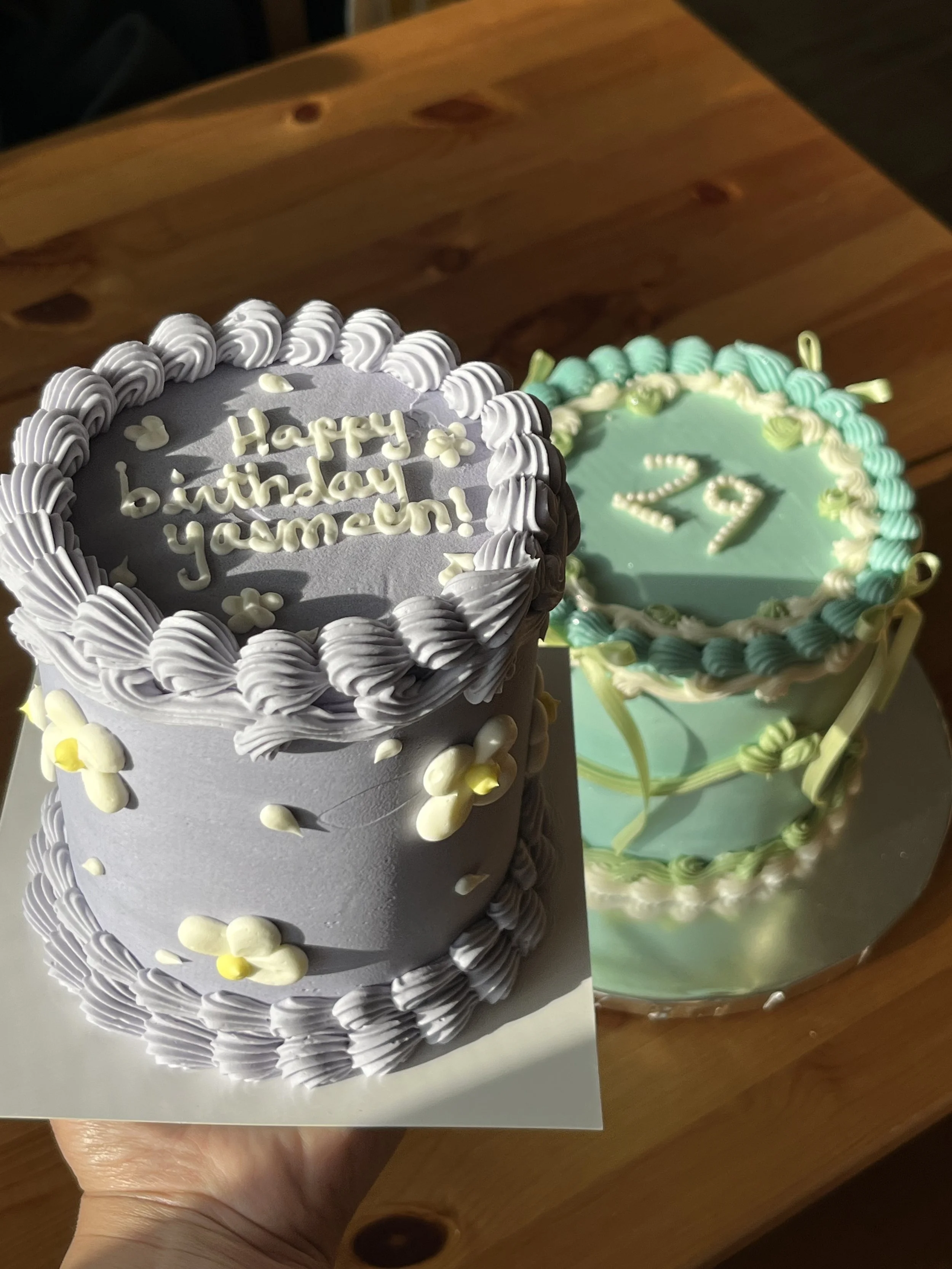 Two decorated birthday cakes, one with purple and white frosting and the message 'Happy birthday yam-zen!', and the other with green and white frosting and the number '29' on top, placed on a wooden surface.