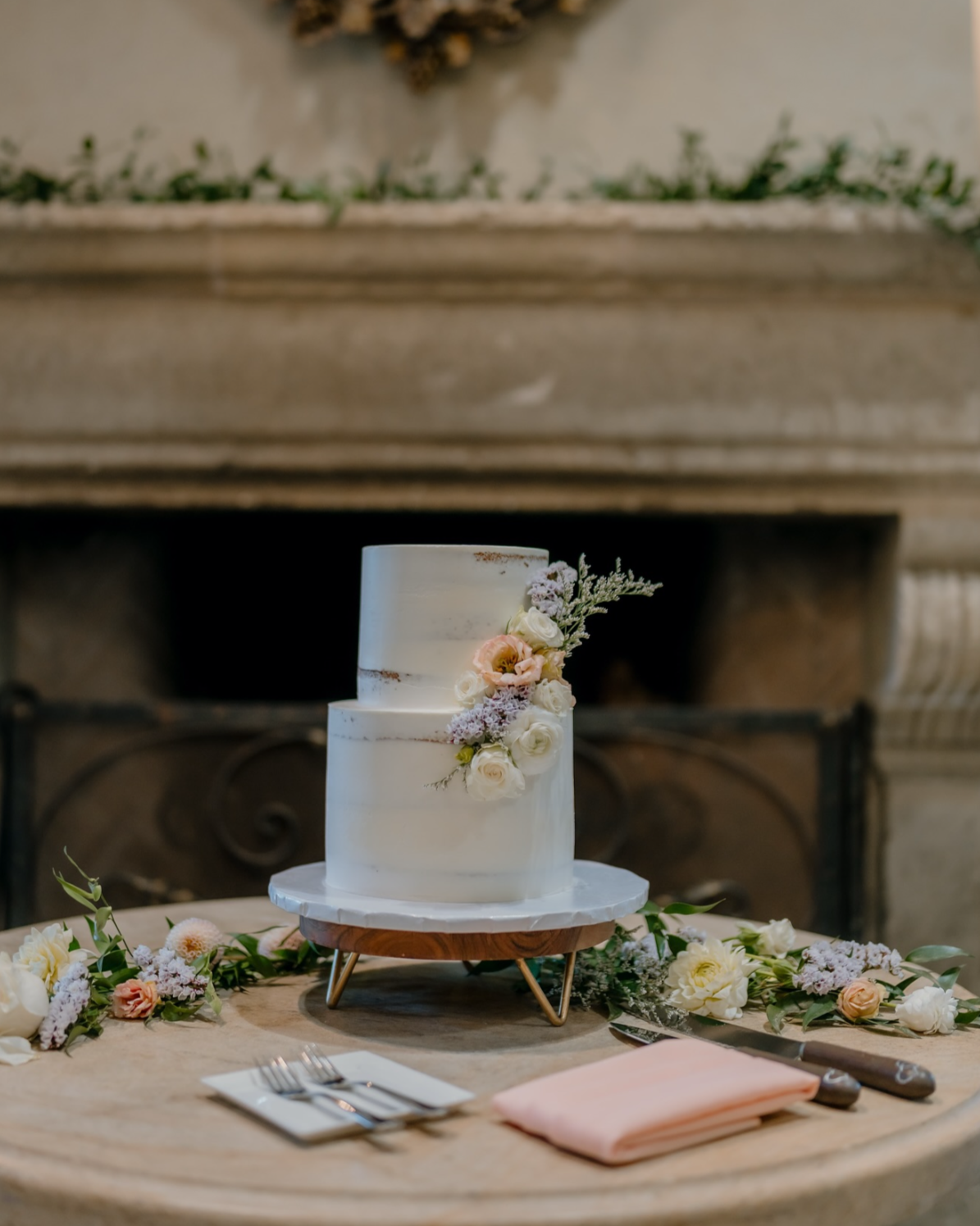 A two-tier white wedding cake decorated with pink and white flowers, placed on a table with floral decorations and surrounded by utensils and napkins.