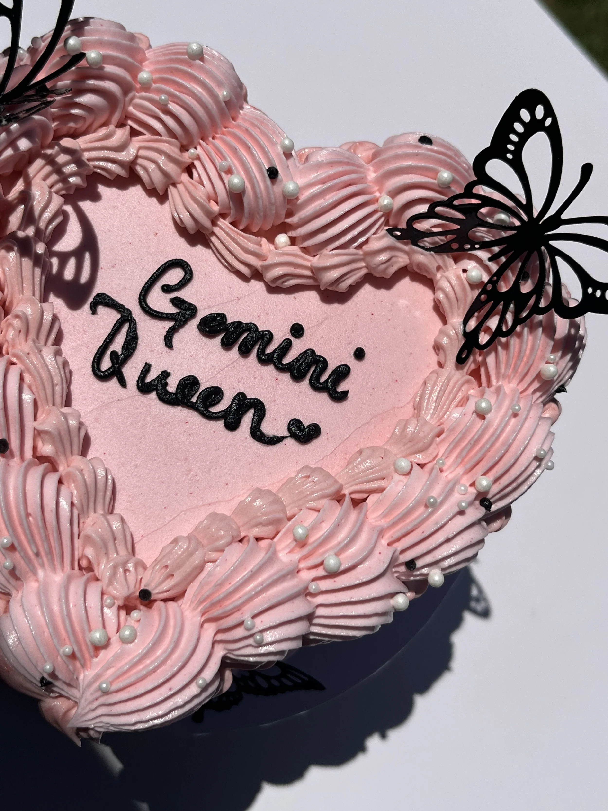Pink heart-shaped cake decorated with pink frosting, white pearl-like sprinkles, and black butterfly cutouts. The cake has a message written in black that says, 'Genie Queen'.