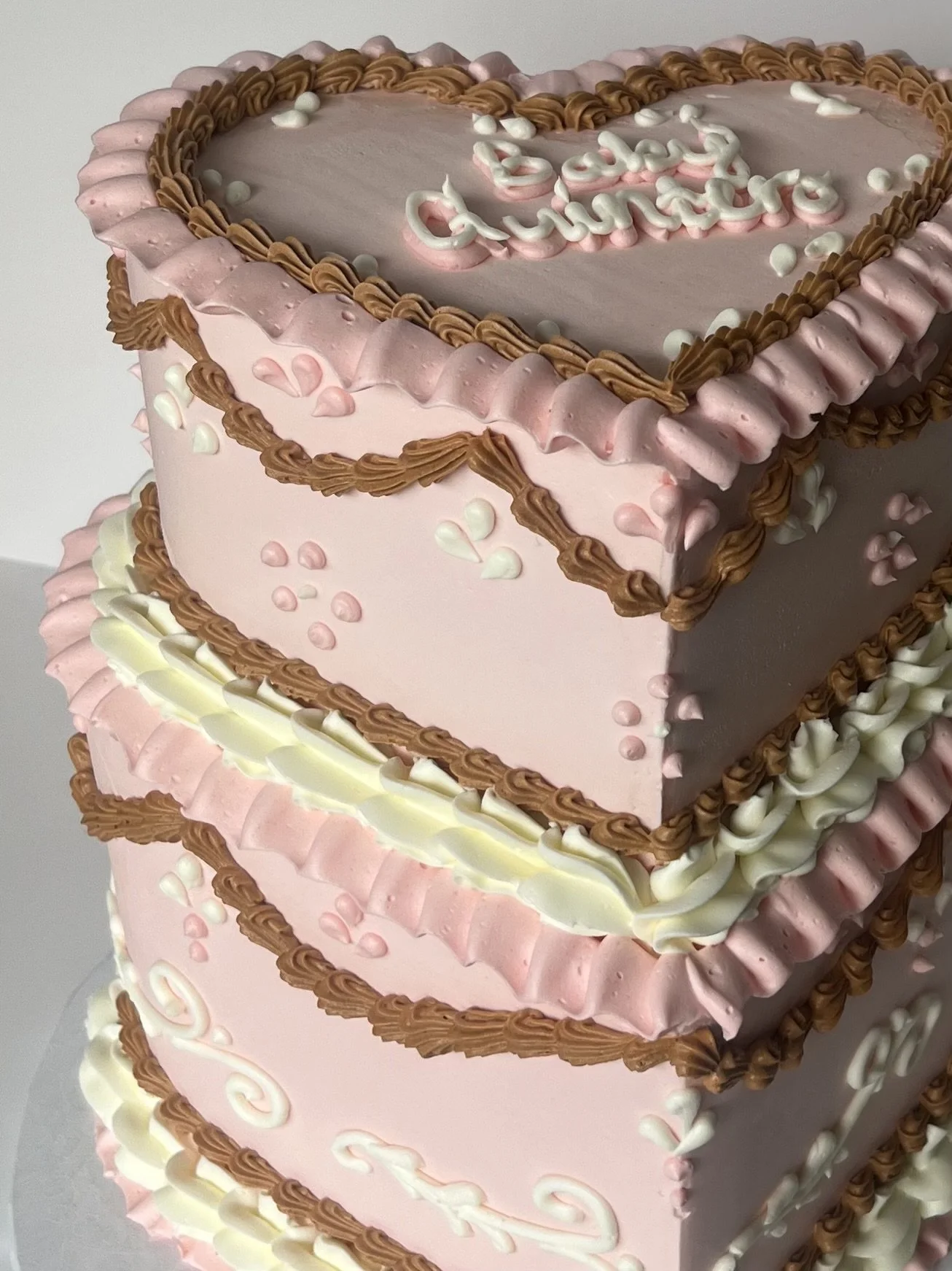 A pink and white multi-tiered cake decorated with icing borders and small hearts, with the words 'Best Grandma' written on the top tier.