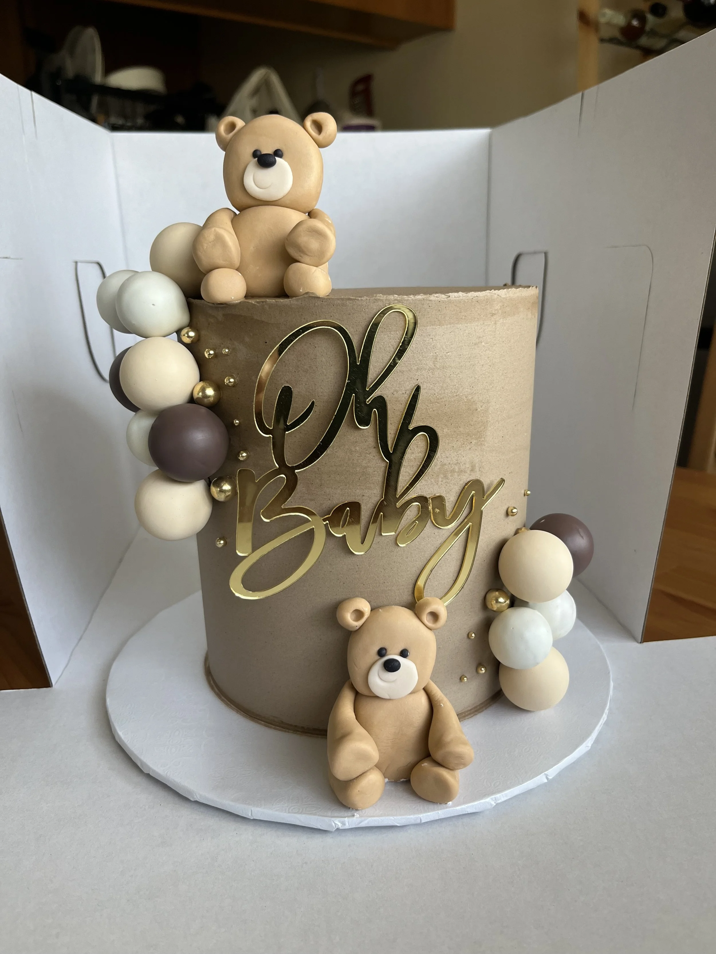A beige and brown teddy bear cake with golden letters, surrounded by cream-colored and purple fondant balls, with a fondant teddy bear on top, displayed on a circular white base.