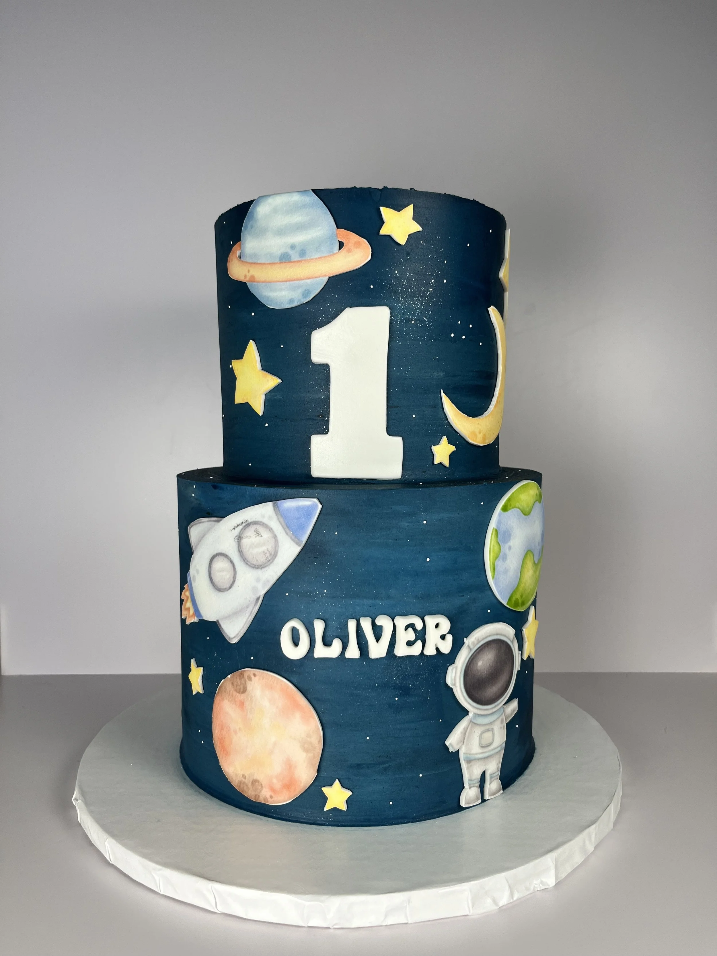 Two-tier space-themed birthday cake with planets, stars, moon, rocket, and astronaut decorations. The name "Oliver" and the number "1" are on the cake, indicating a first birthday celebration.