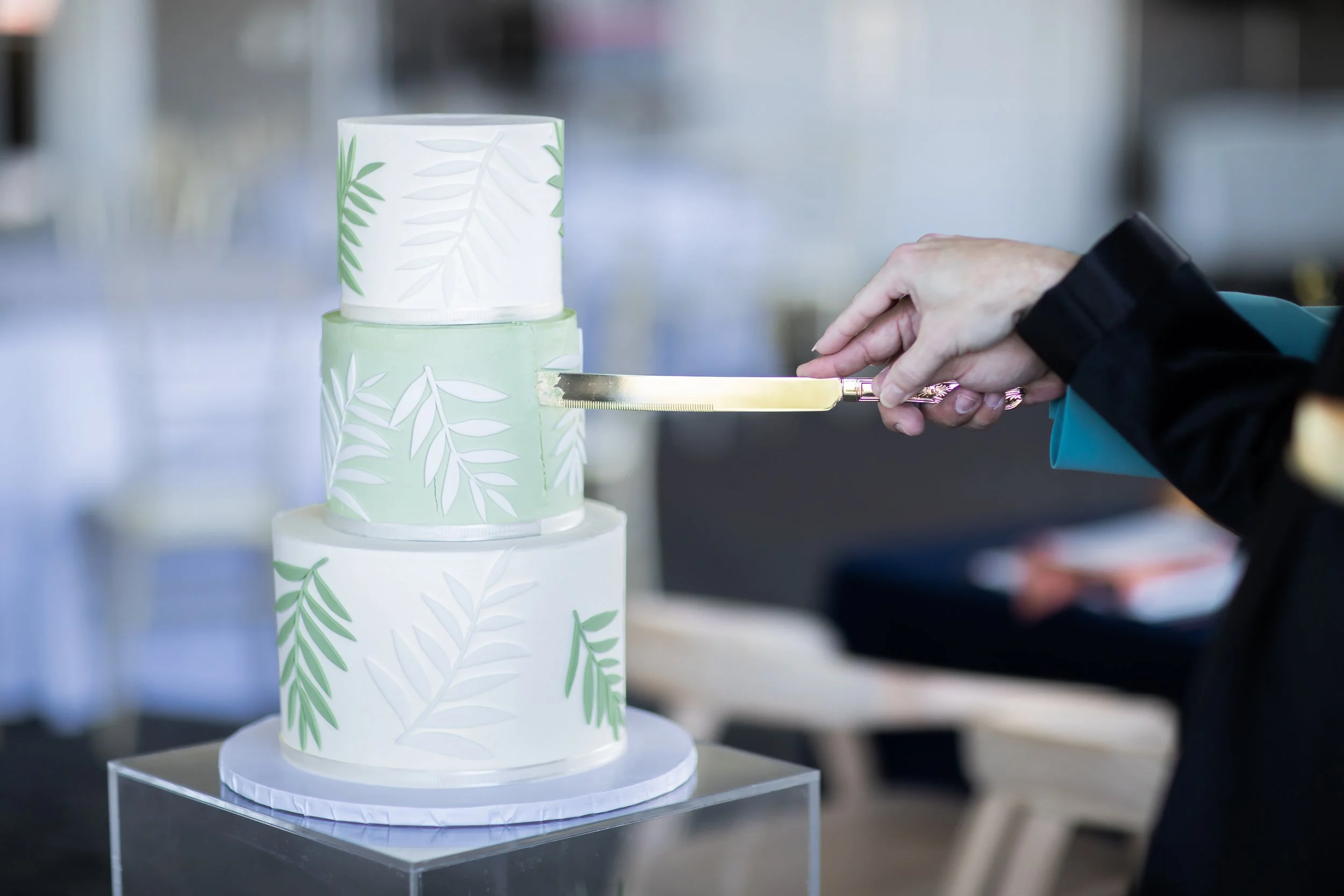 A multi-tiered wedding cake decorated with white and light green fondant, featuring a leaf pattern. A person is slicing the cake with a gold-colored knife.