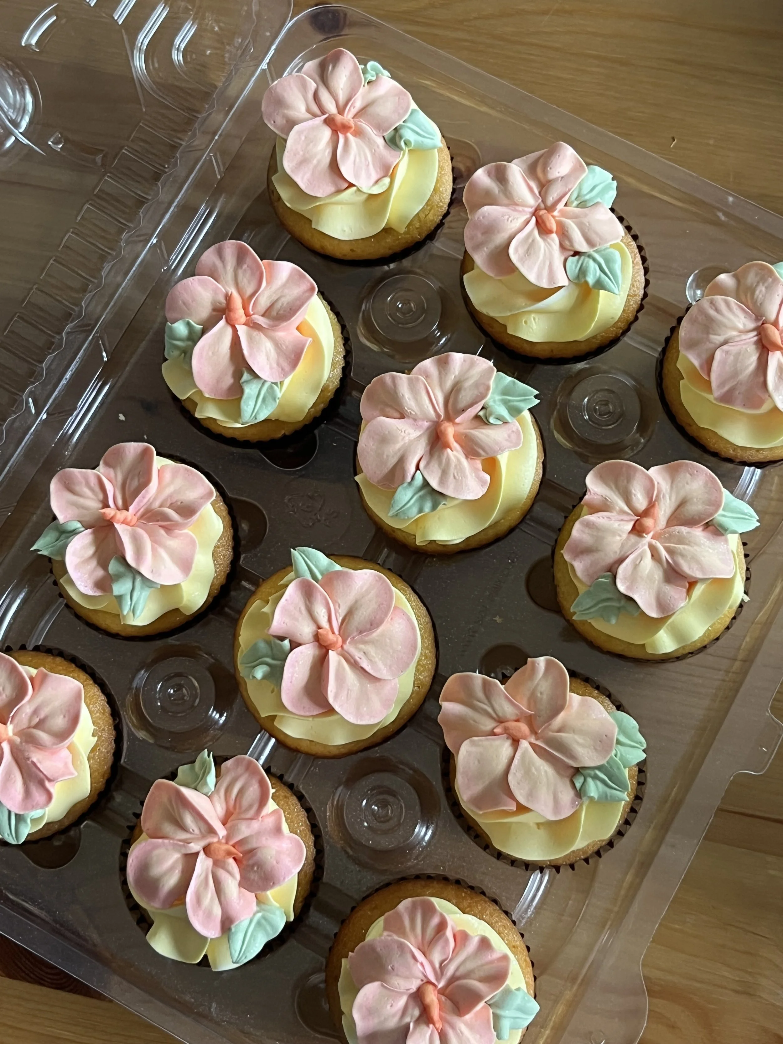 Tray of cupcakes decorated with pastel pink, yellow, and green frosting, topped with large flower-shaped decorations.