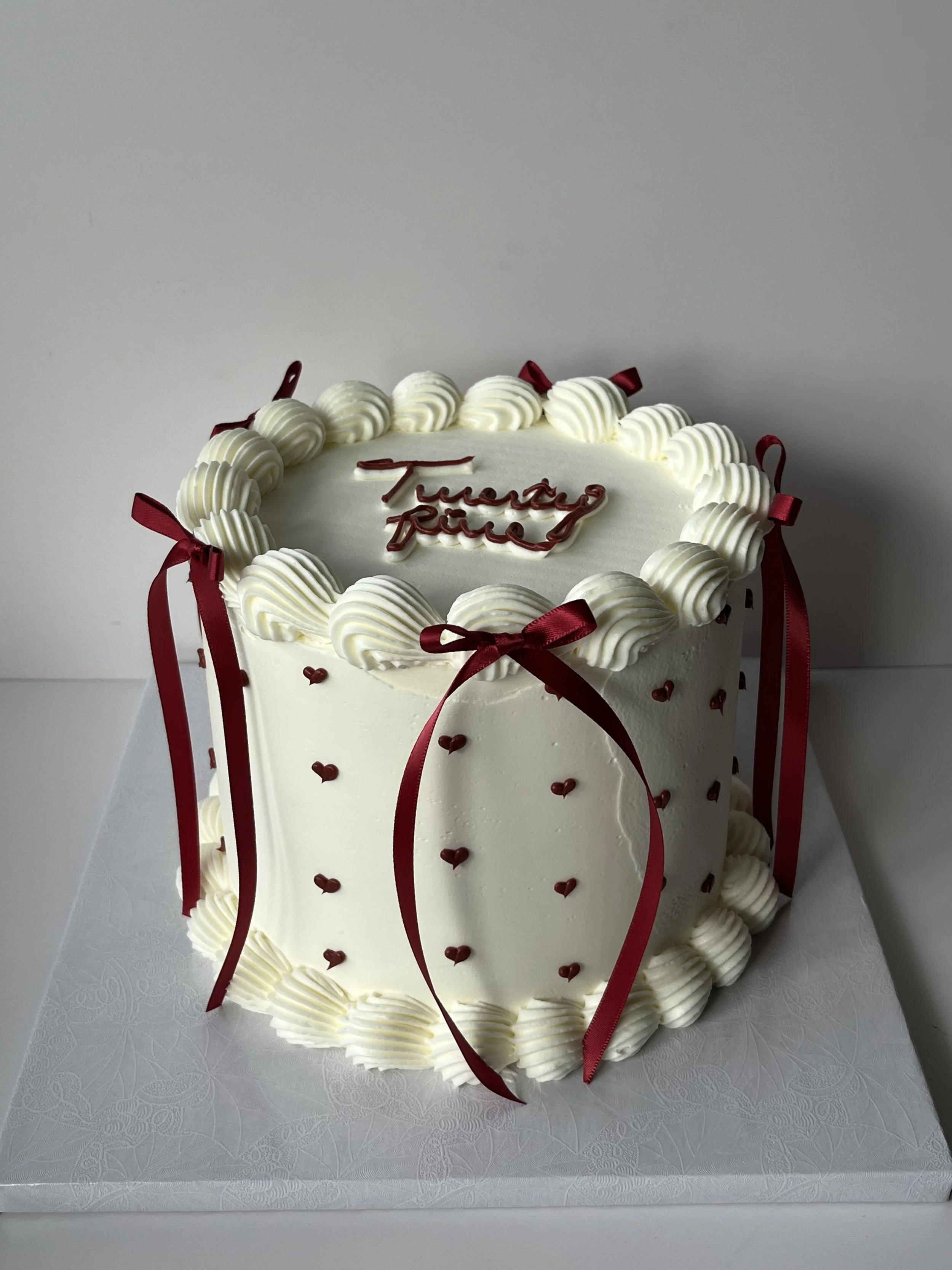 A white, heart-shaped cake decorated with white buttercream swirls and red ribbons, featuring small red hearts, and a message in red icing that says 'Treasure Dad' on top.