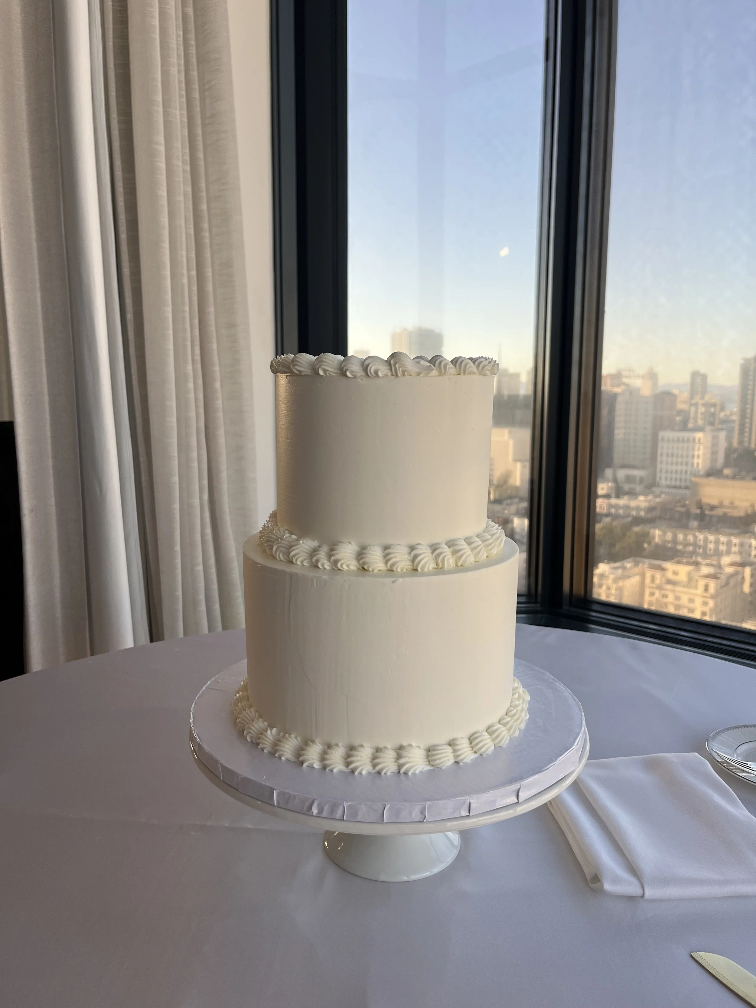 Two-tiered white wedding cake with piped decorative borders, placed on a table near a large window with city skyscrapers in the background.