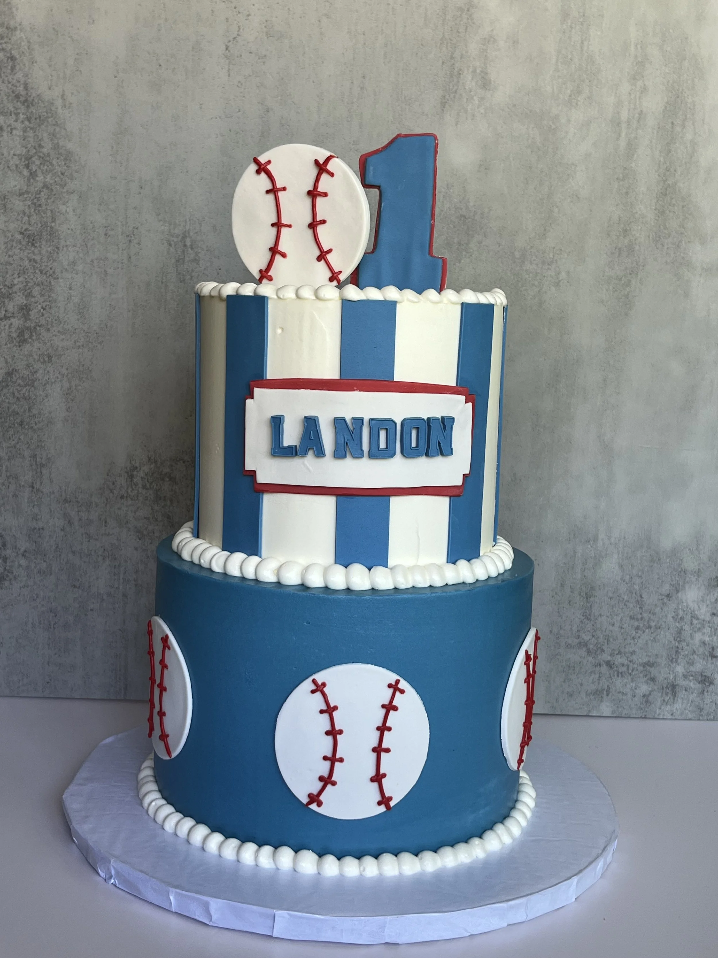A tiered birthday cake decorated with a baseball theme. The cake has blue and white vertical stripes, baseballs with red stitches, a large blue number 1 on top, and a white oval with the name 'Landon' in blue letters. The cake is bordered with white 
