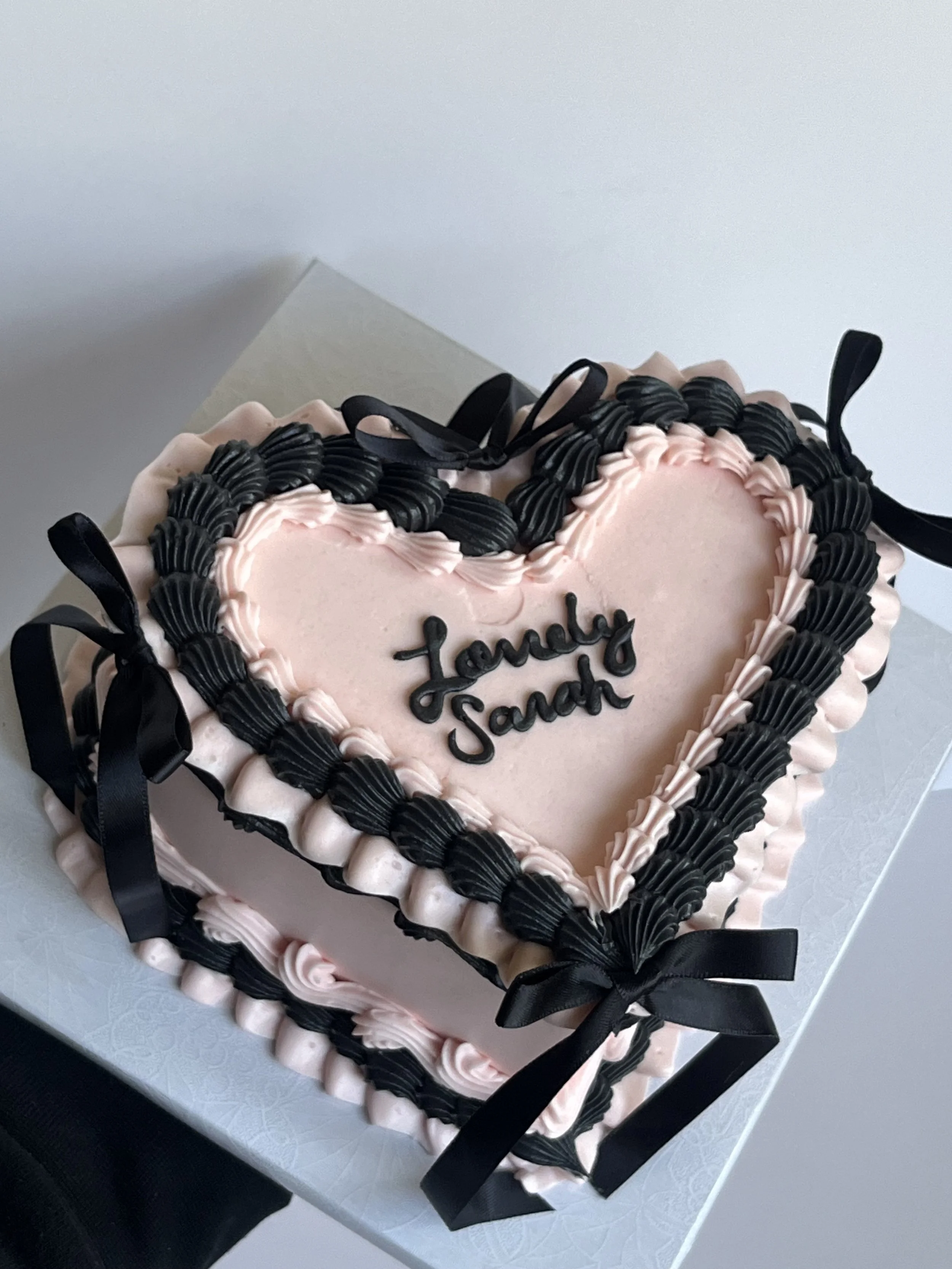 Heart-shaped birthday cake with pink and black icing, decorated with black ribbon bows and words 'Lena & Sandh' written in black icing.