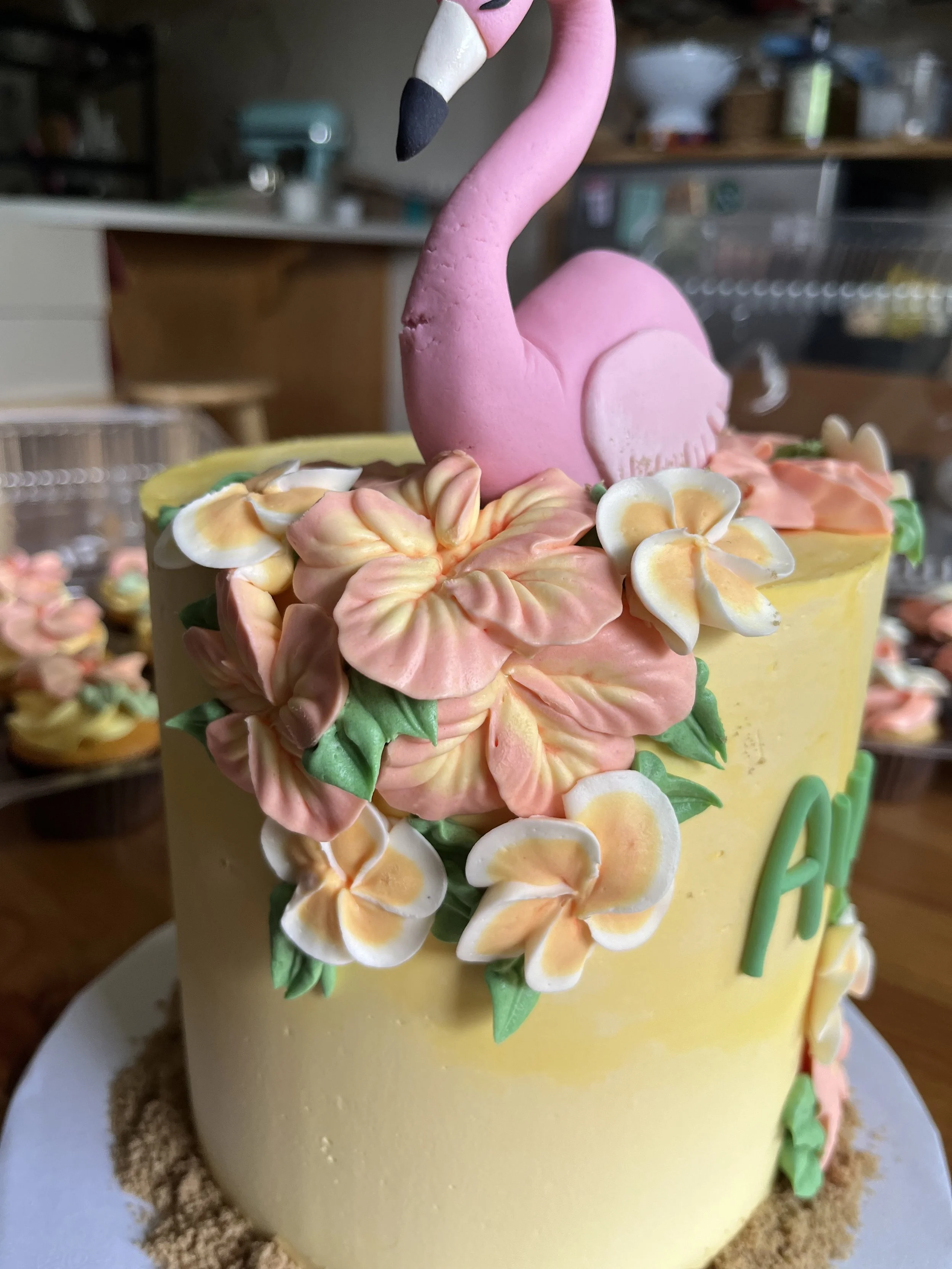 A cake decorated with a pink flamingo figure on top, surrounded by pastel-colored flowers and green leaves, with a yellow exterior and a sandy-colored base.