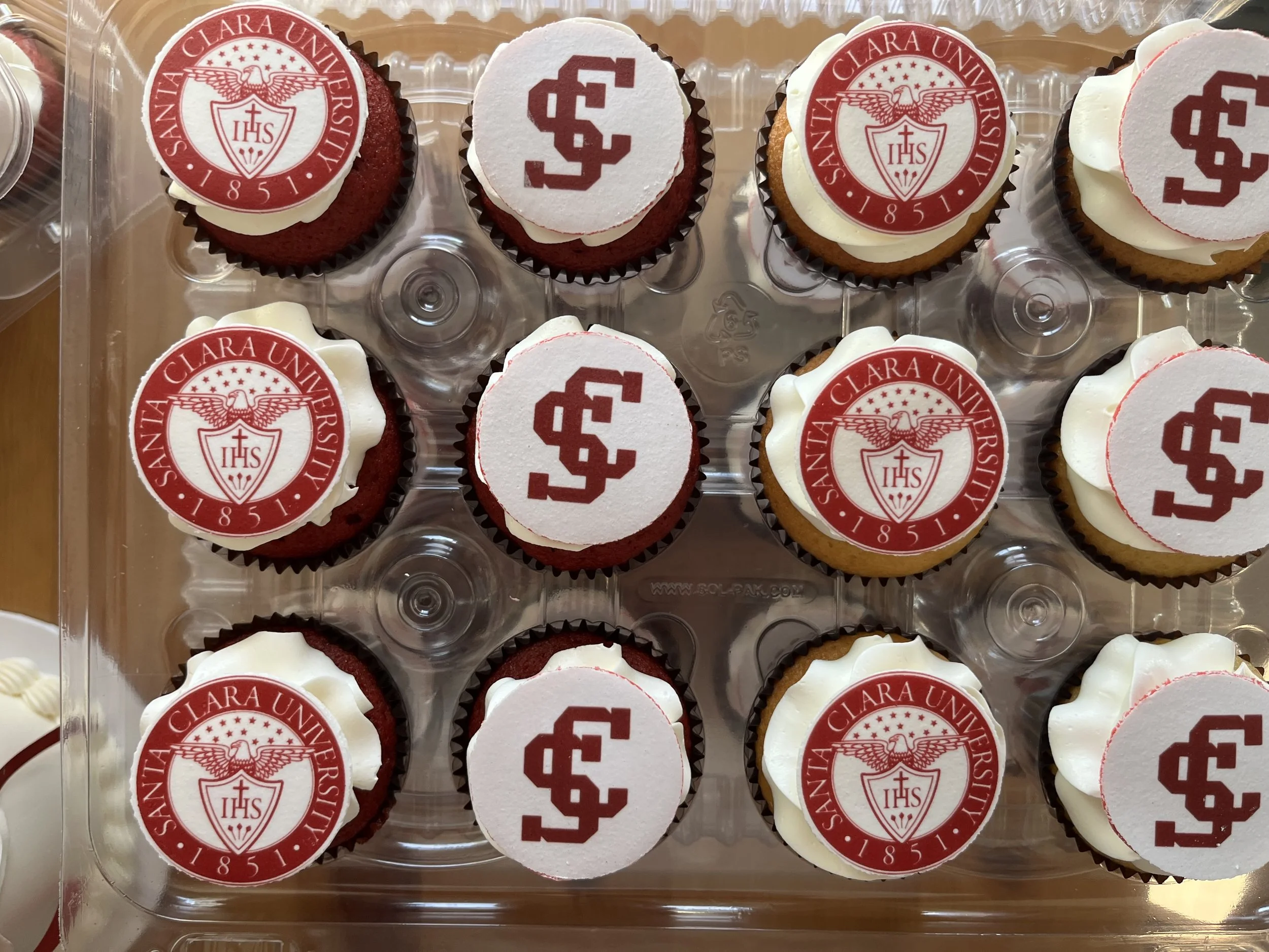 A plastic container of cupcakes featuring red and white edible toppers with the San Clara University crest and initials 'SC'.