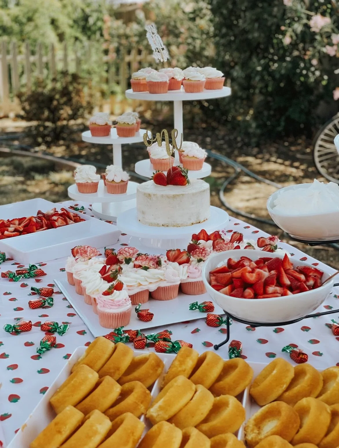 Table with strawberry-themed desserts, including cupcakes with white frosting, a white cake with strawberries and a cake topper, bowls of sliced strawberries, pink frosted cupcakes, and fried treats, set outdoors with greenery in the background
