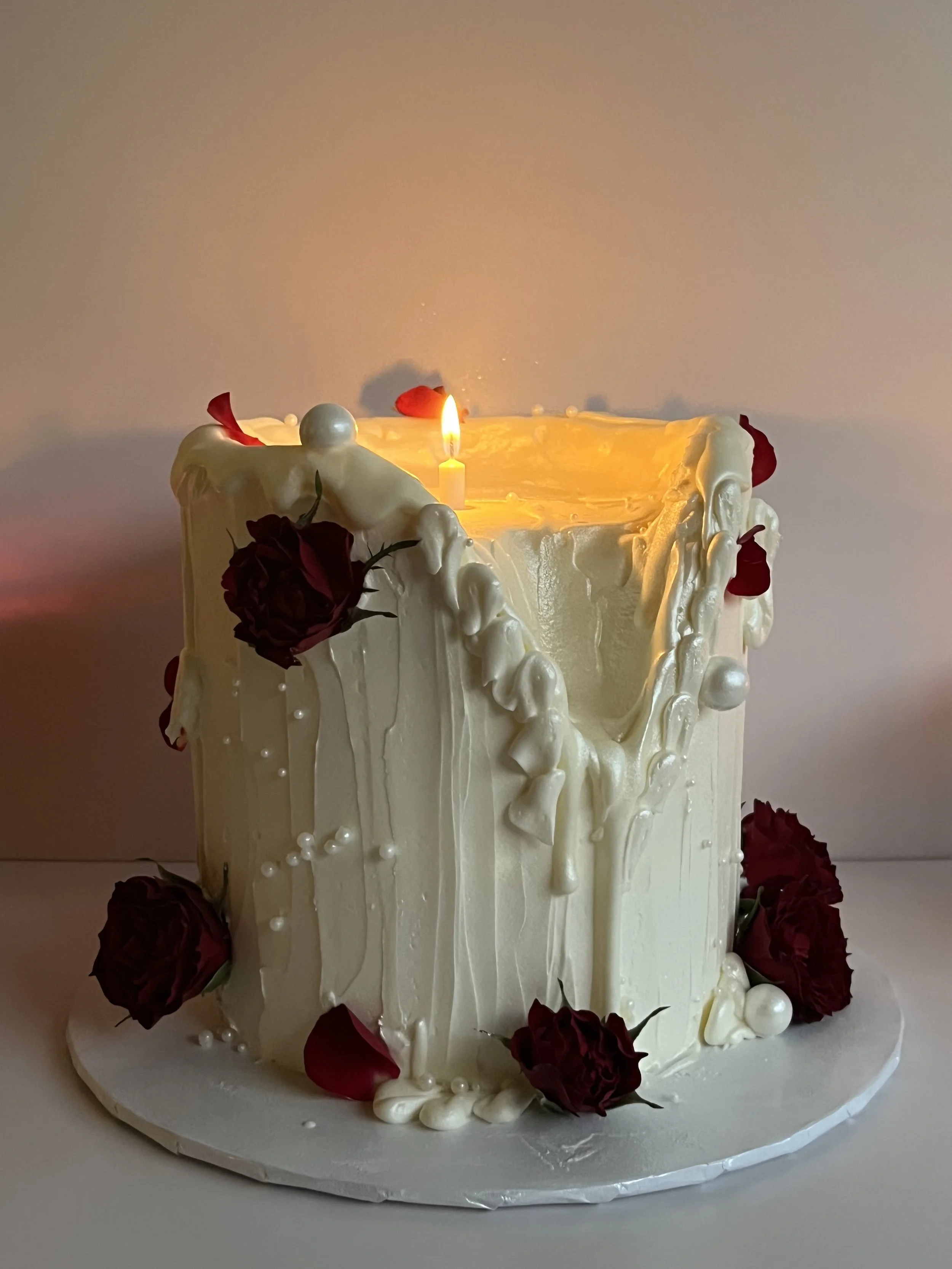 A tall white wedding cake decorated with red roses and white dragees, with a lit candle at the top center.