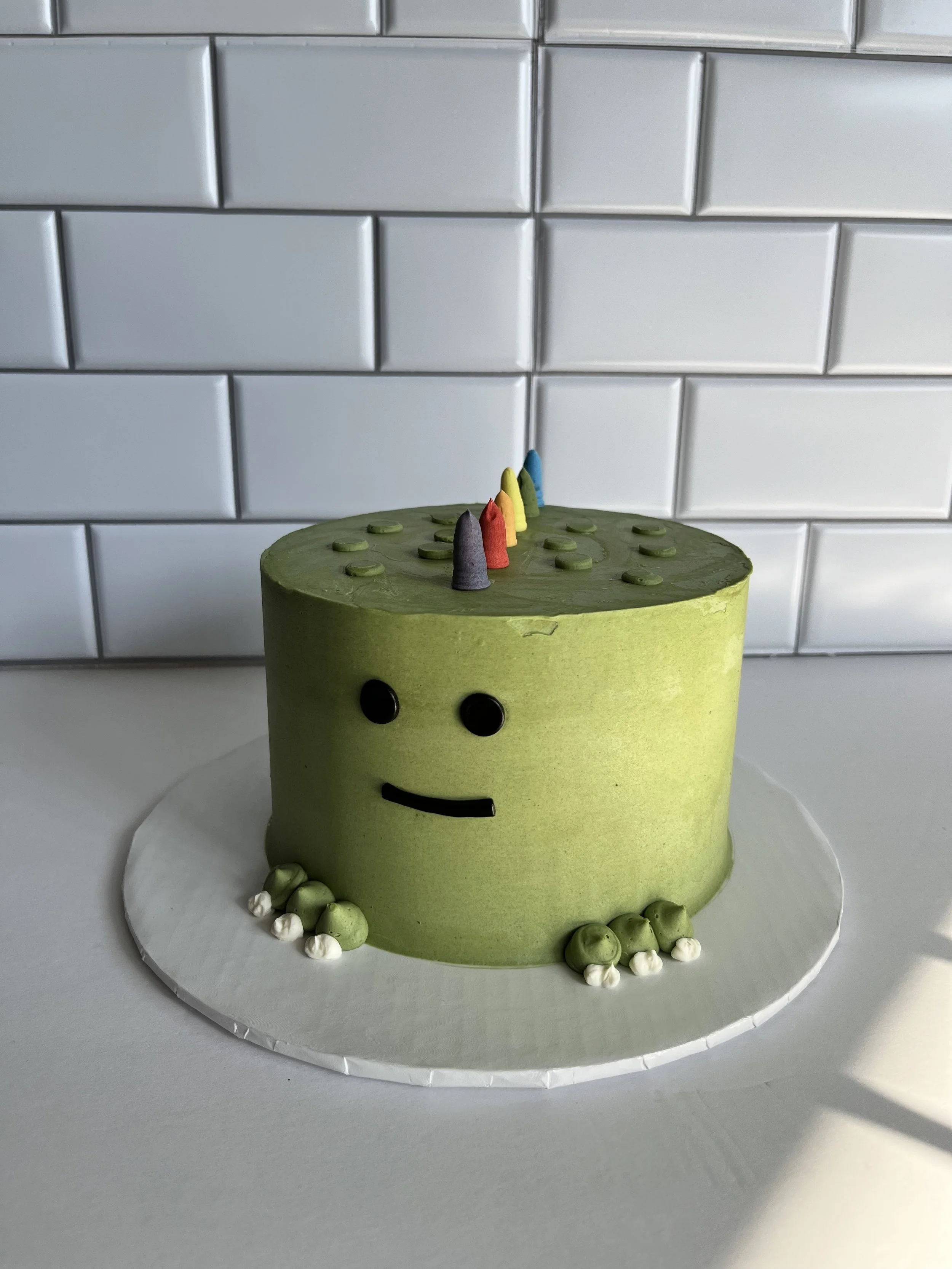 A green, cylindrical cake with a happy face, topped with small colorful candles and surrounded by green and white balls, placed on a round white cake board.