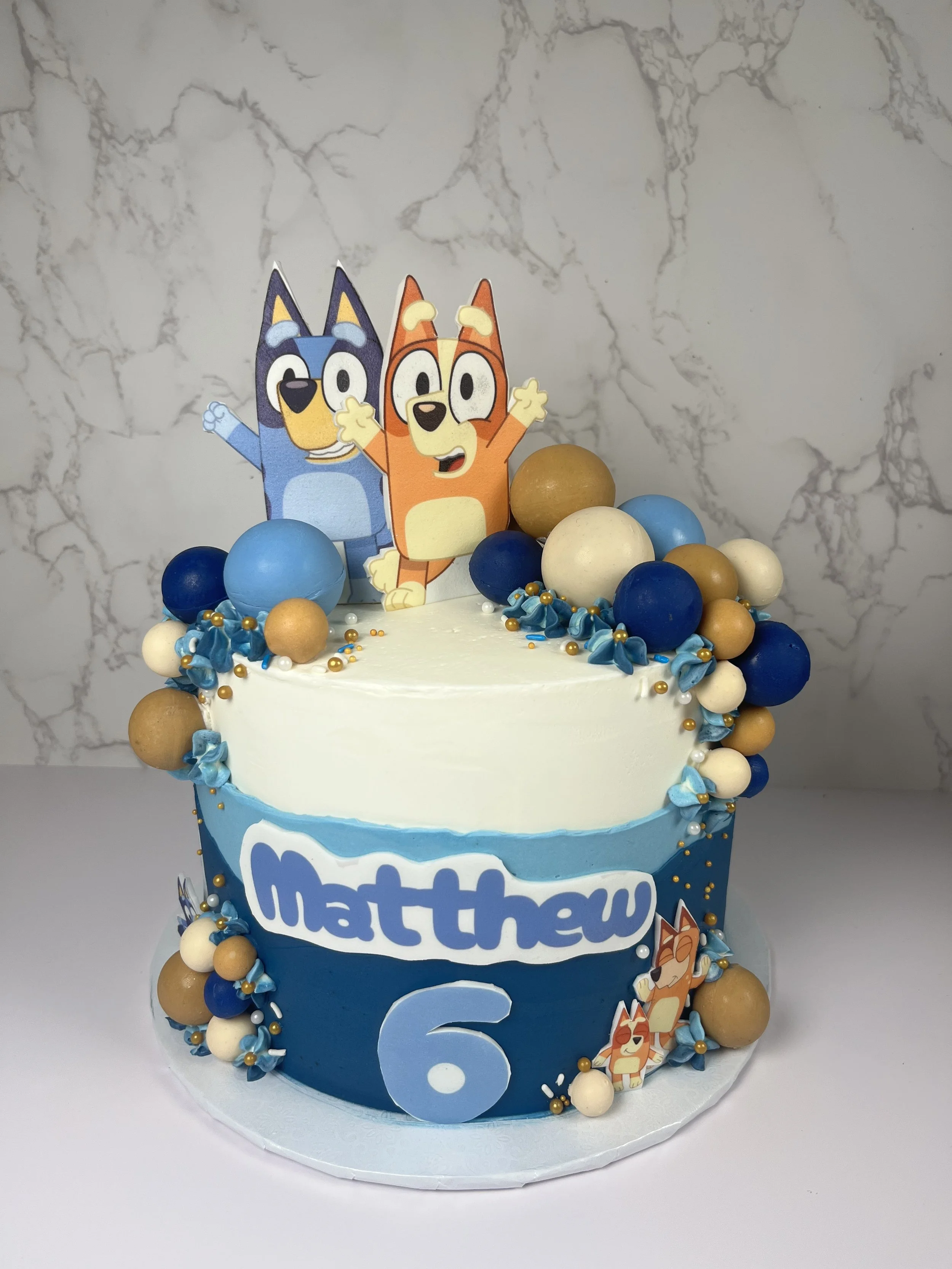 A birthday cake decorated with cartoon animal toppers and blue, white, and gold spheres. The cake has blue and white icing and a large blue number 6 on the front. The background is a marble wall.
