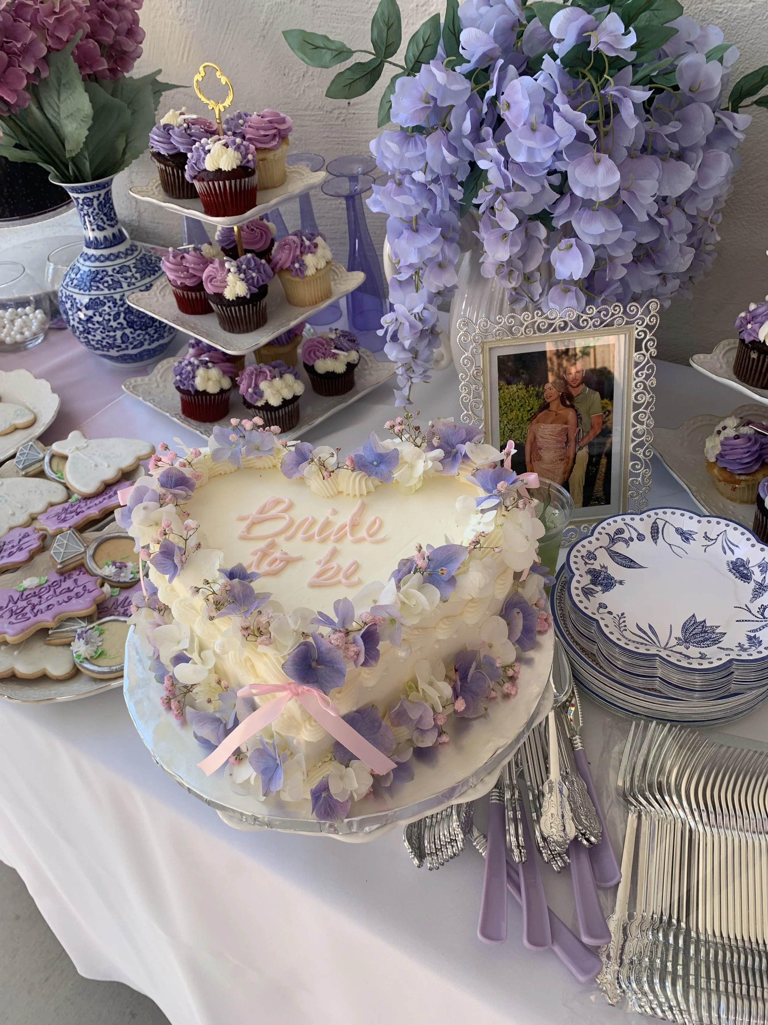 A dessert table with a large white cake decorated with purple and white flowers, a tiered cupcake stand with purple, white, and chocolate cupcakes, purple utensils, floral arrangements in purple and white, a framed photo of a couple, and decorative p