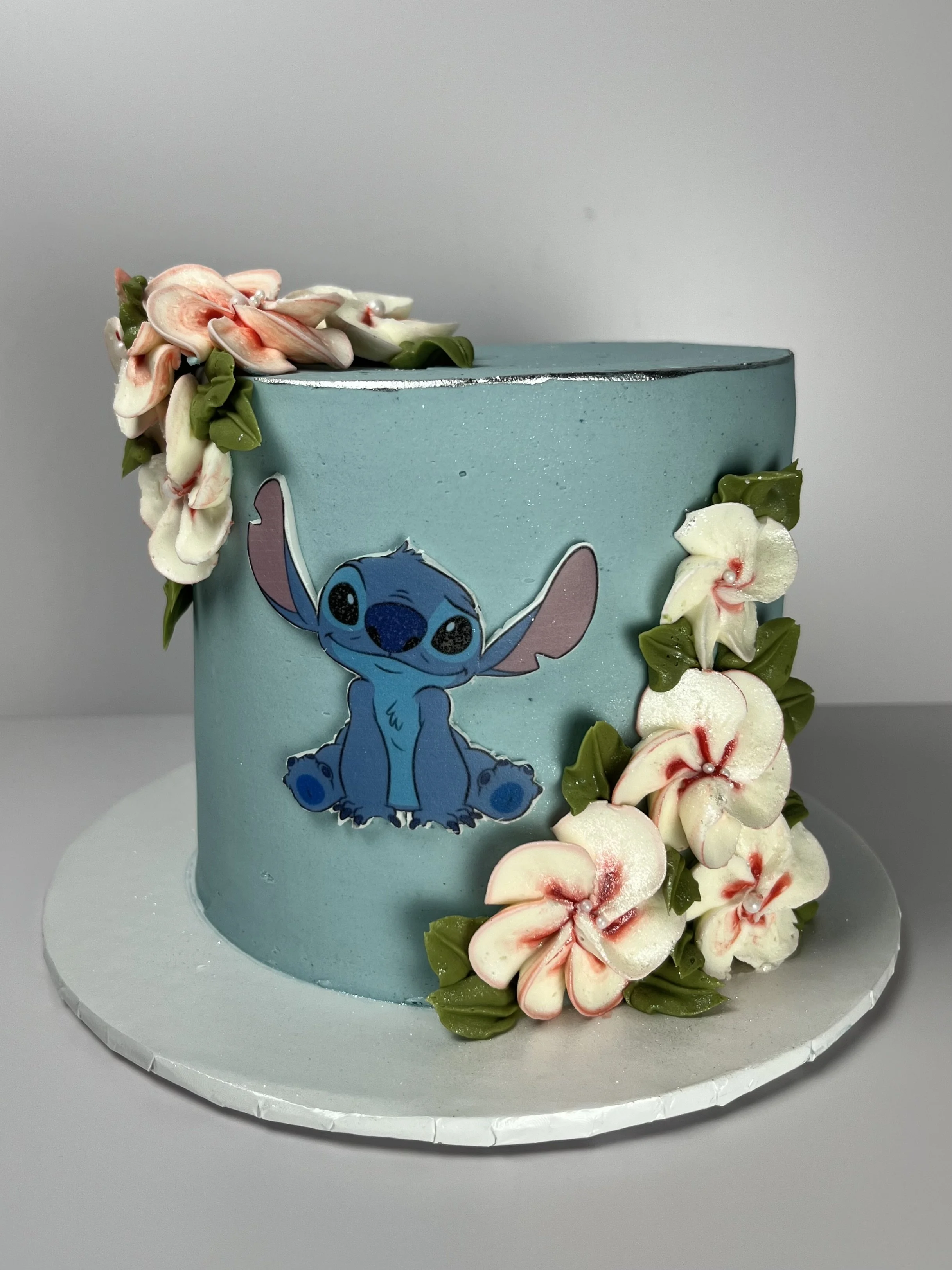 A light blue cake decorated with white and pink flower-shaped decorations cascading down the side and a cartoon character of Stitch from Disney's 'Lilo & Stitch' on the front.