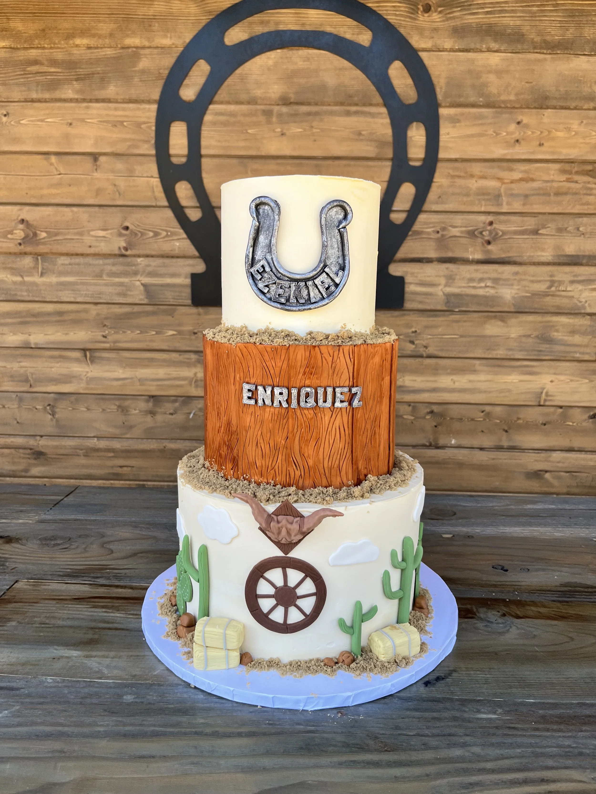 A multi-tiered cake with a cowboy and western theme, decorated with cactus, hay bales, and a wagon wheel, topped with a white candle featuring a horseshoe and the name 'Enriquez'.