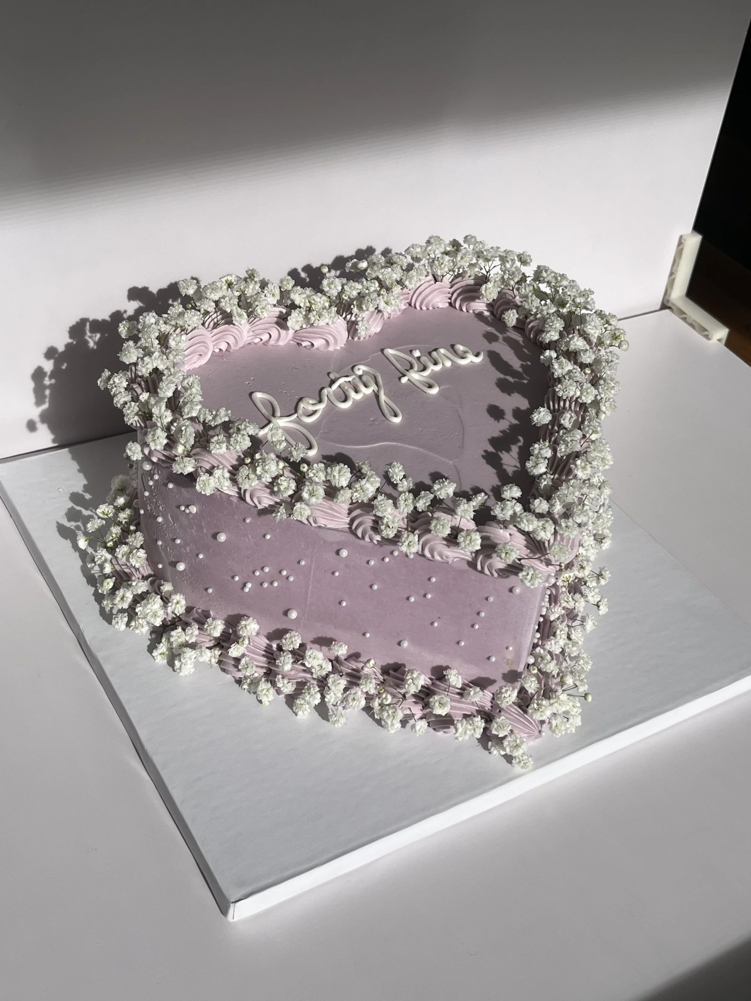 A heart-shaped pink birthday cake decorated with white flowers and pink icing piping, with the words "Loving You" written on top.