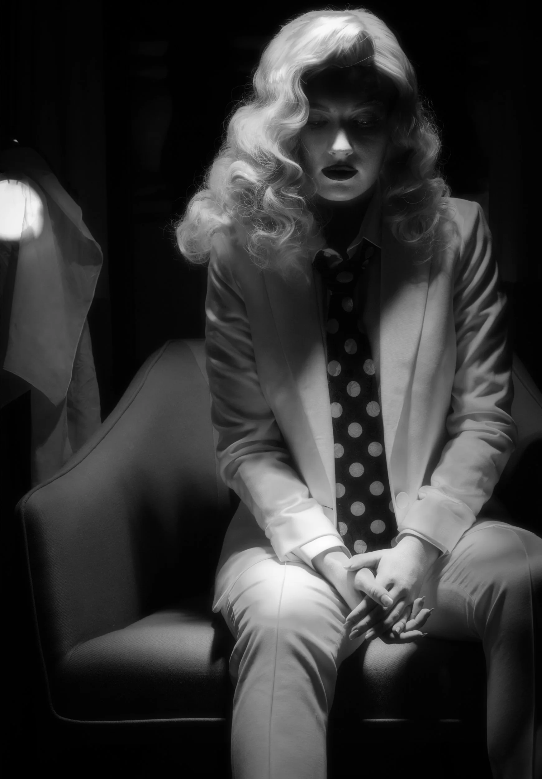 Black-and-white portrait of a woman with wavy blonde hair sitting in a chair, wearing a light blazer and polka-dot tie against a dark background.