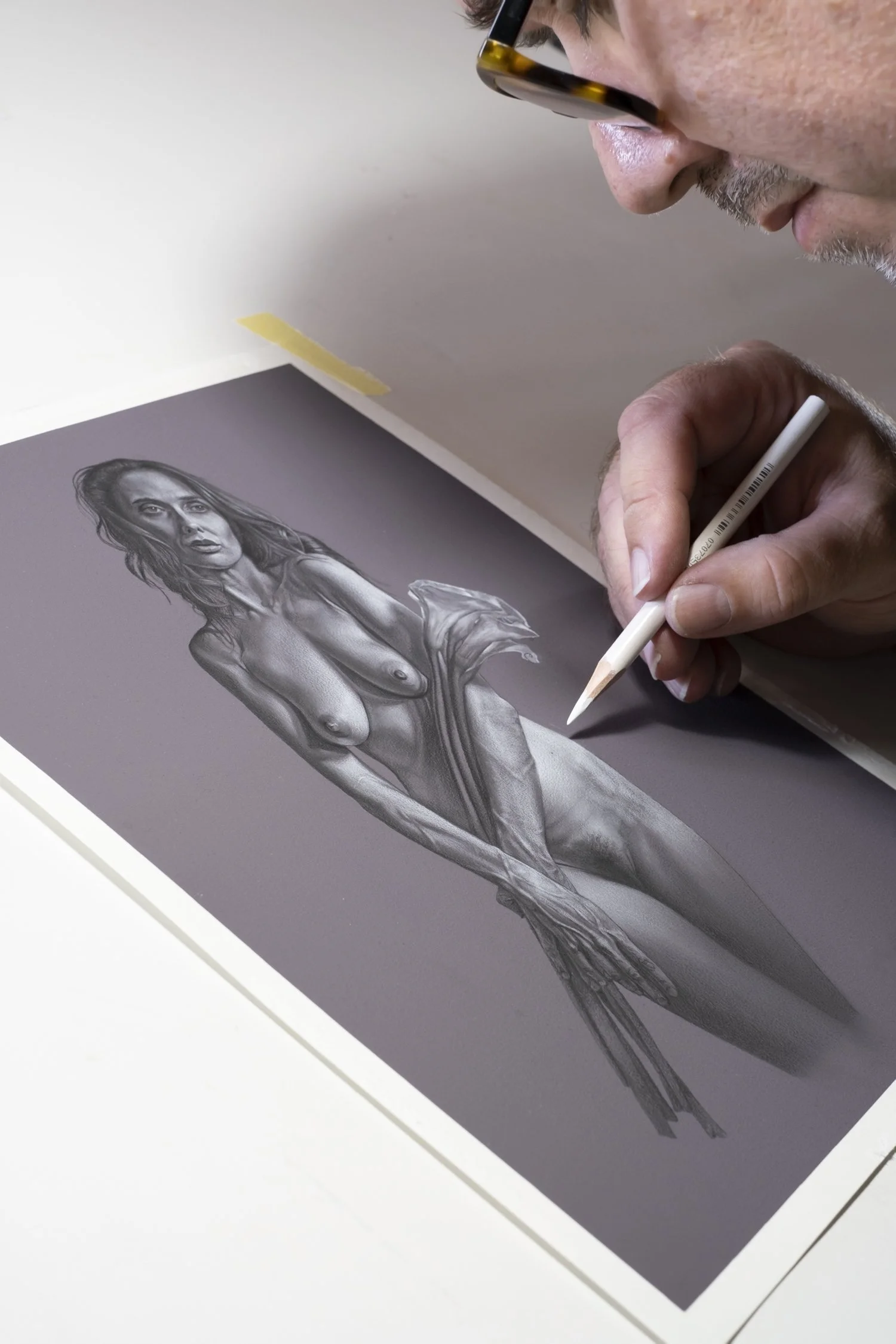 Artist wearing glasses draws a detailed graphite portrait of a nude woman with a white pencil.