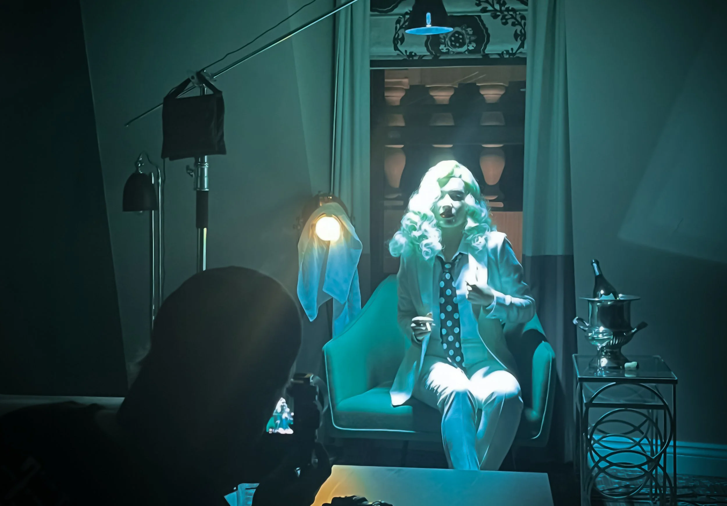 Person in white clown makeup and curly wig seated in an armchair, holding a phone in a dim blue-lit room