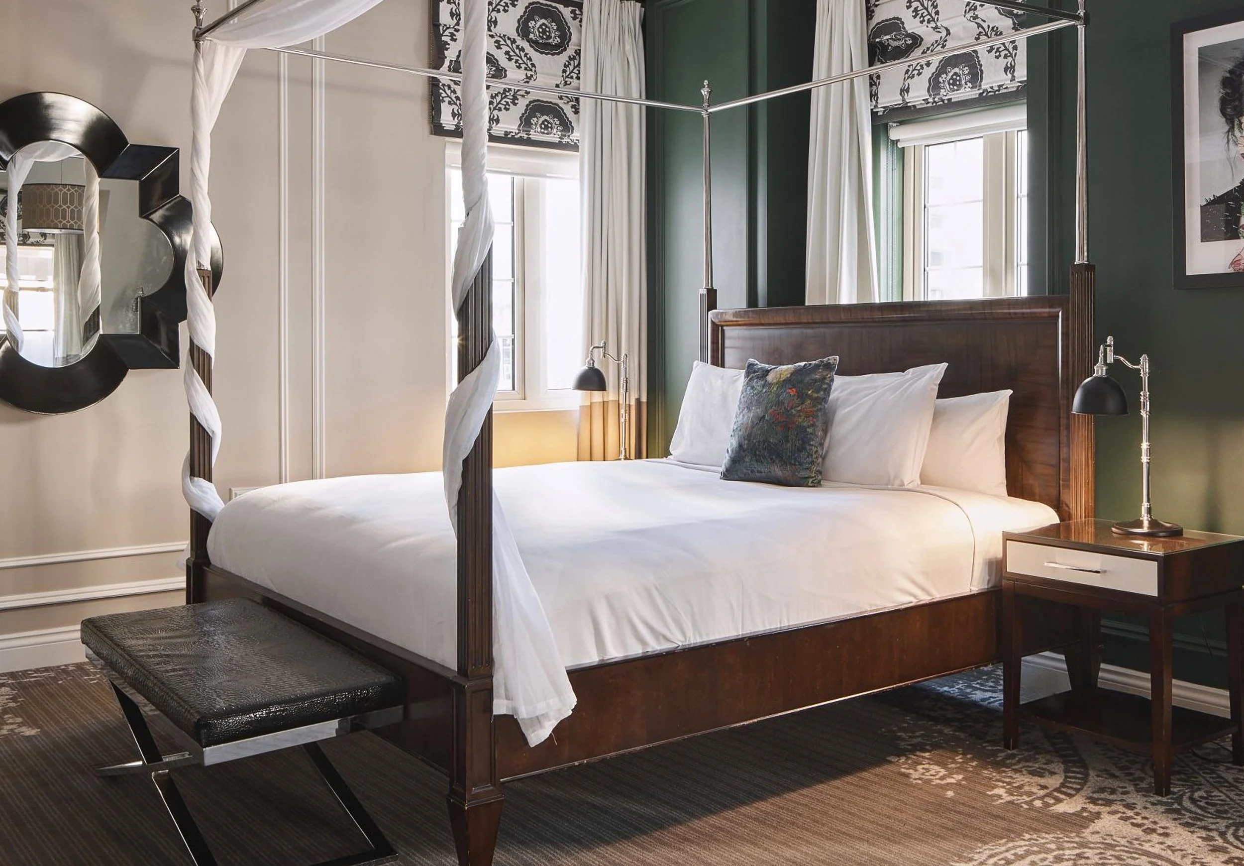 Elegant bedroom with a dark wood canopy bed, white linens, patterned curtains, bedside lamps, and a large wall mirror