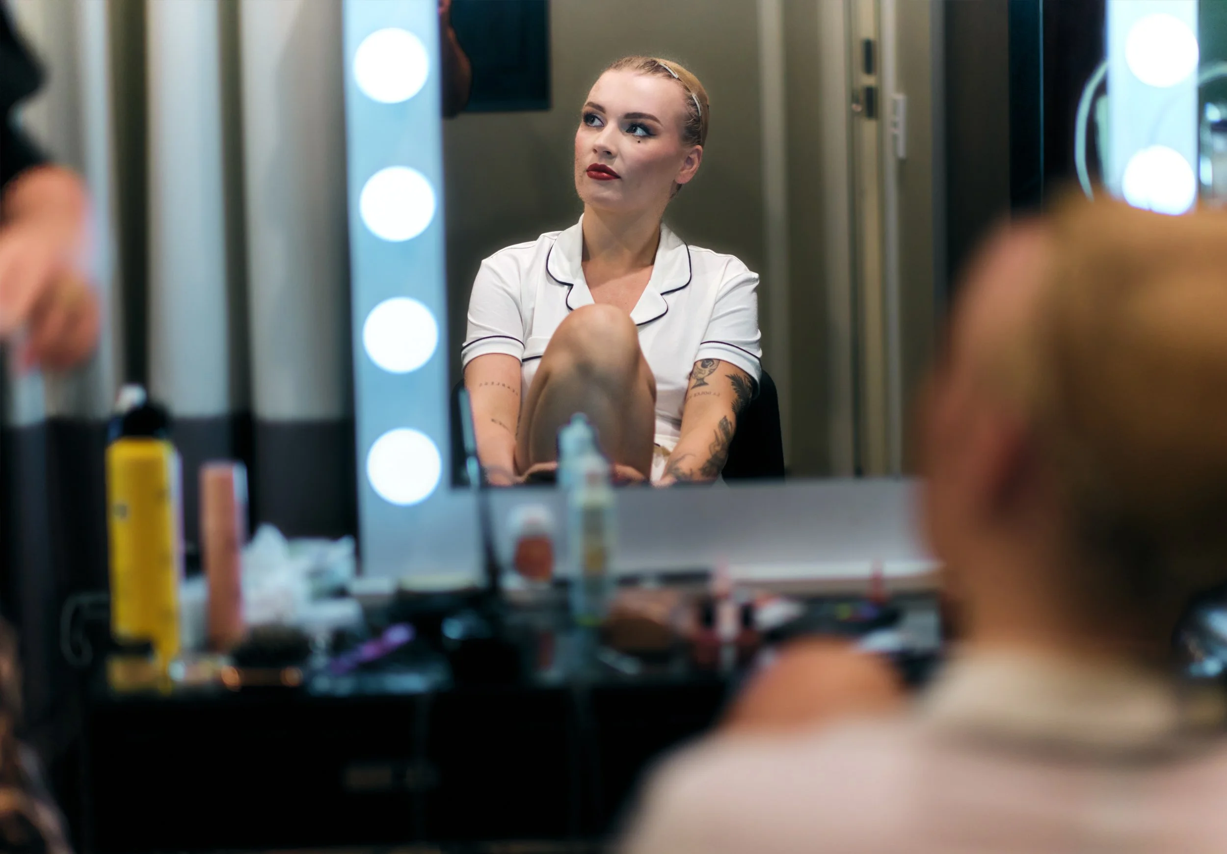 Tattooed woman in a white shirt sitting in a makeup room, reflected in a mirror surrounded by vanity lights and cosmetics.