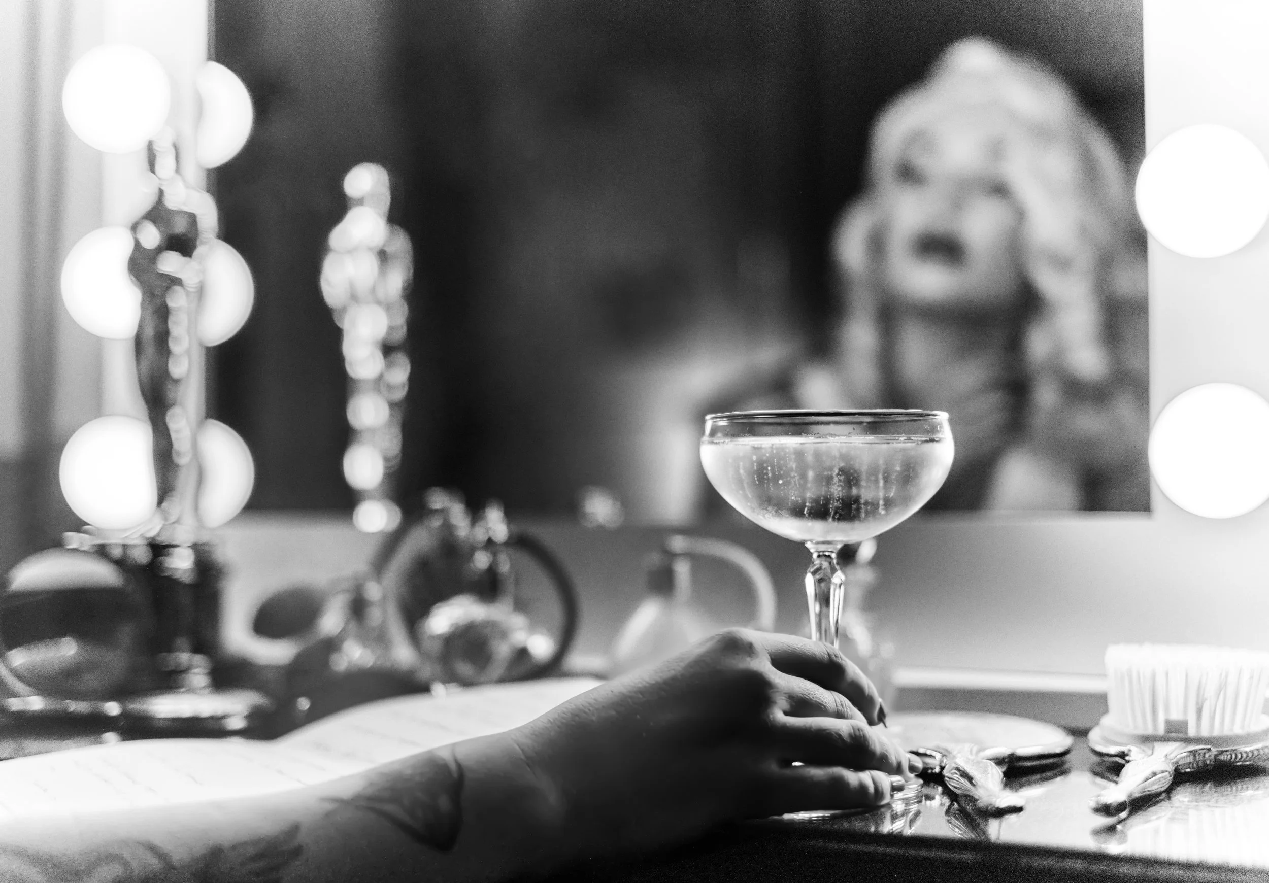 Black-and-white restaurant scene with a person’s hand beside a coupe glass, tableware, and a blurred diner in the background