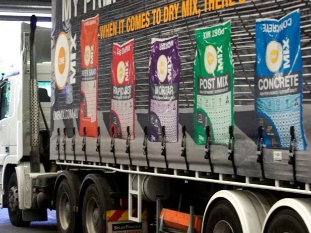 A truck displaying large bags of concrete mix in red, purple, green, and blue, each labeled for different types of concrete including rapid set, mortar, post mix, and concrete.