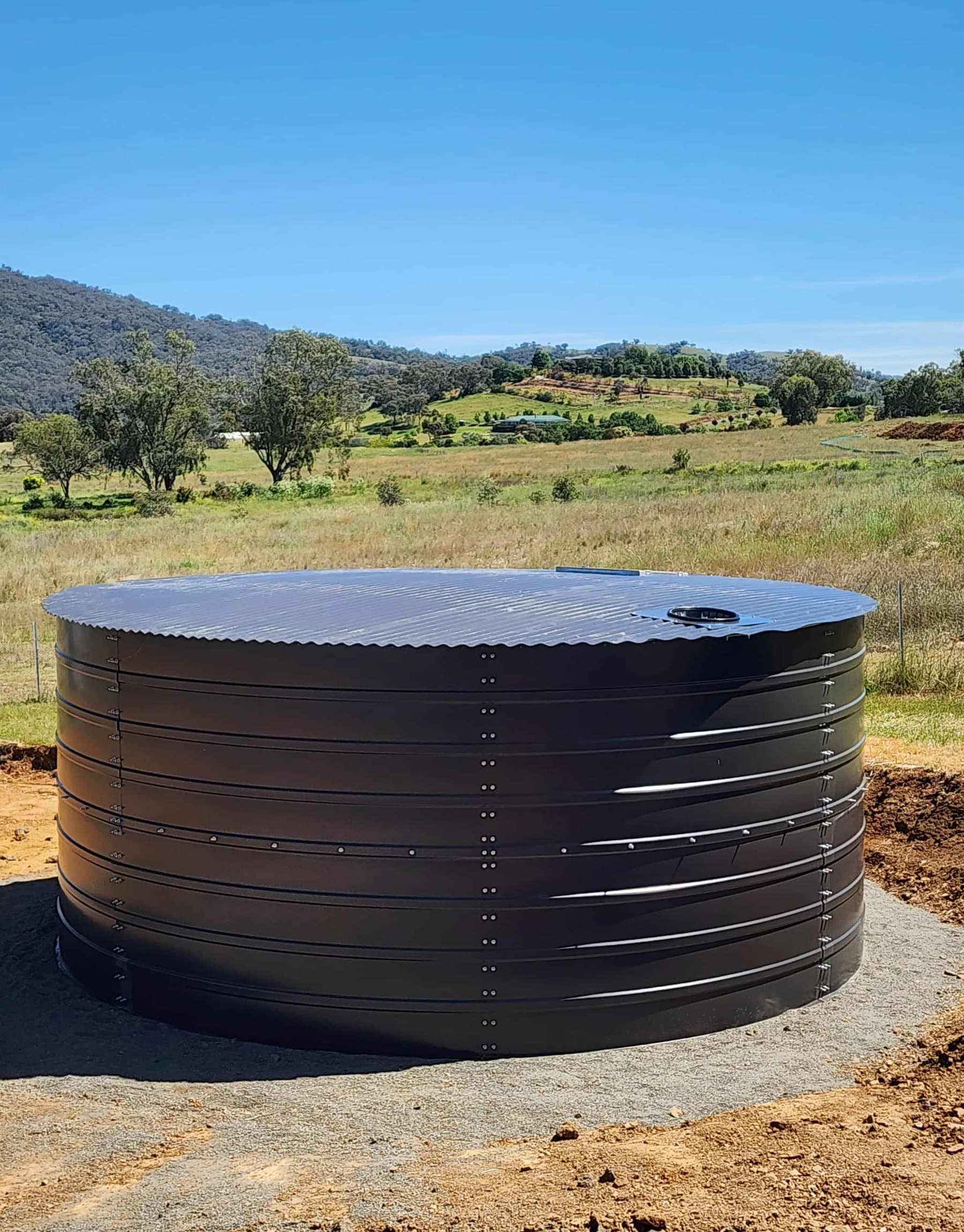 Large black rainwater tank in a rural landscape with hills, trees, and fields under a clear blue sky.