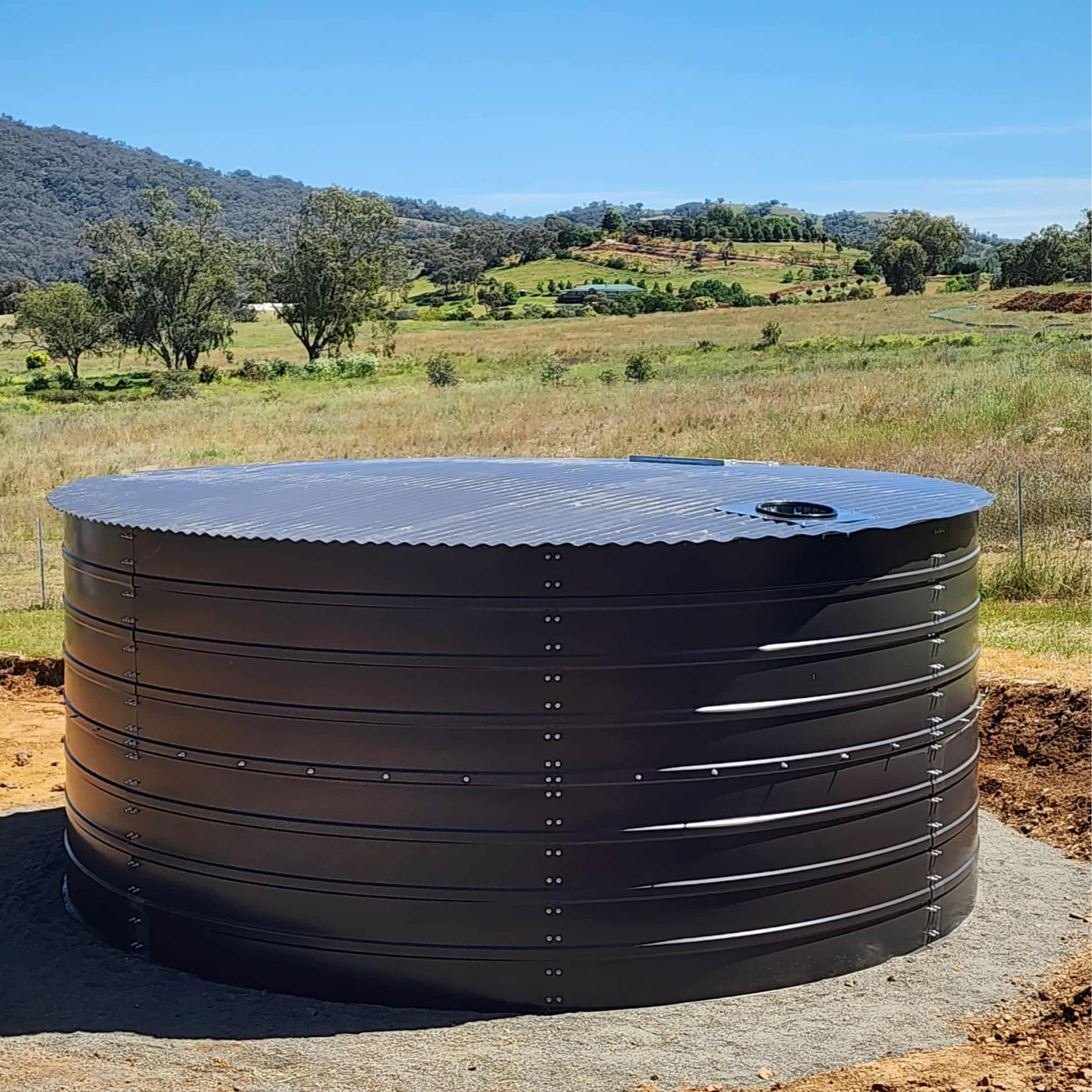 Large black water storage tank in a rural landscape with rolling hills and scattered trees under a clear blue sky.