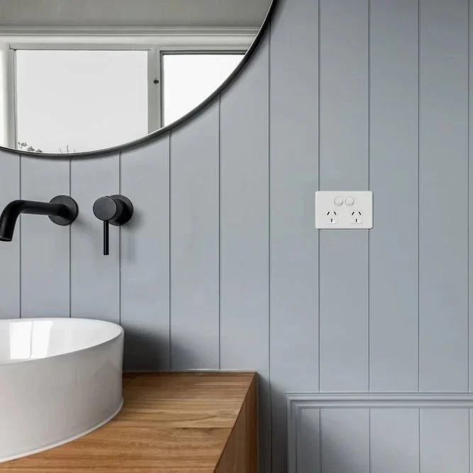 Bathroom vanity with a white vessel sink, black wall-mounted faucet, wooden countertop, a round mirror, gray shiplap wall, electrical outlet, and a window reflecting trees outside.