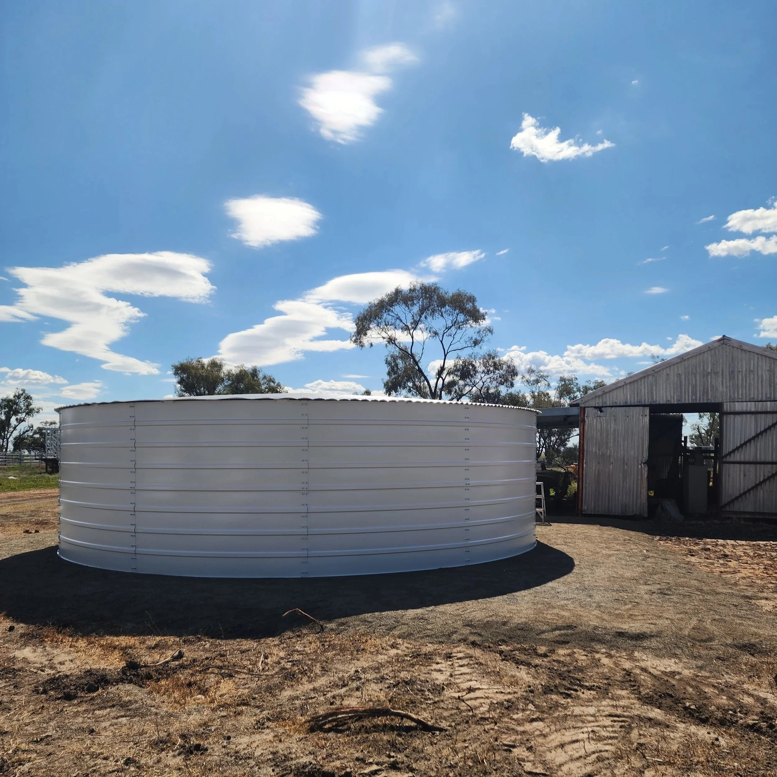 A clear blue sky with scattered white clouds over a rural landscape with trees, a large water tank, and a metal building.